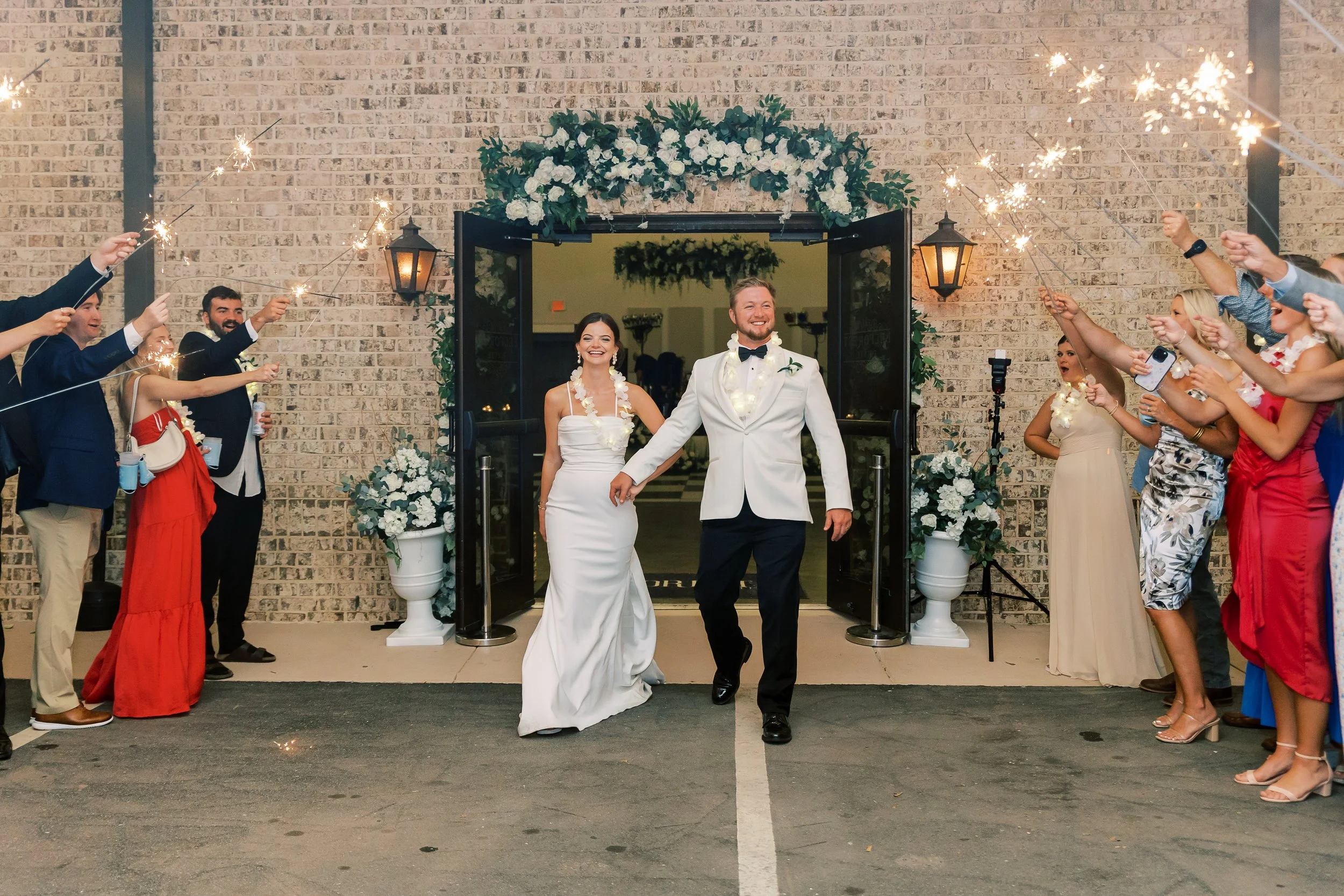 Bride and groom walking out of a wedding venue, smiling, with guests on both sides holding sparklers, celebrating.