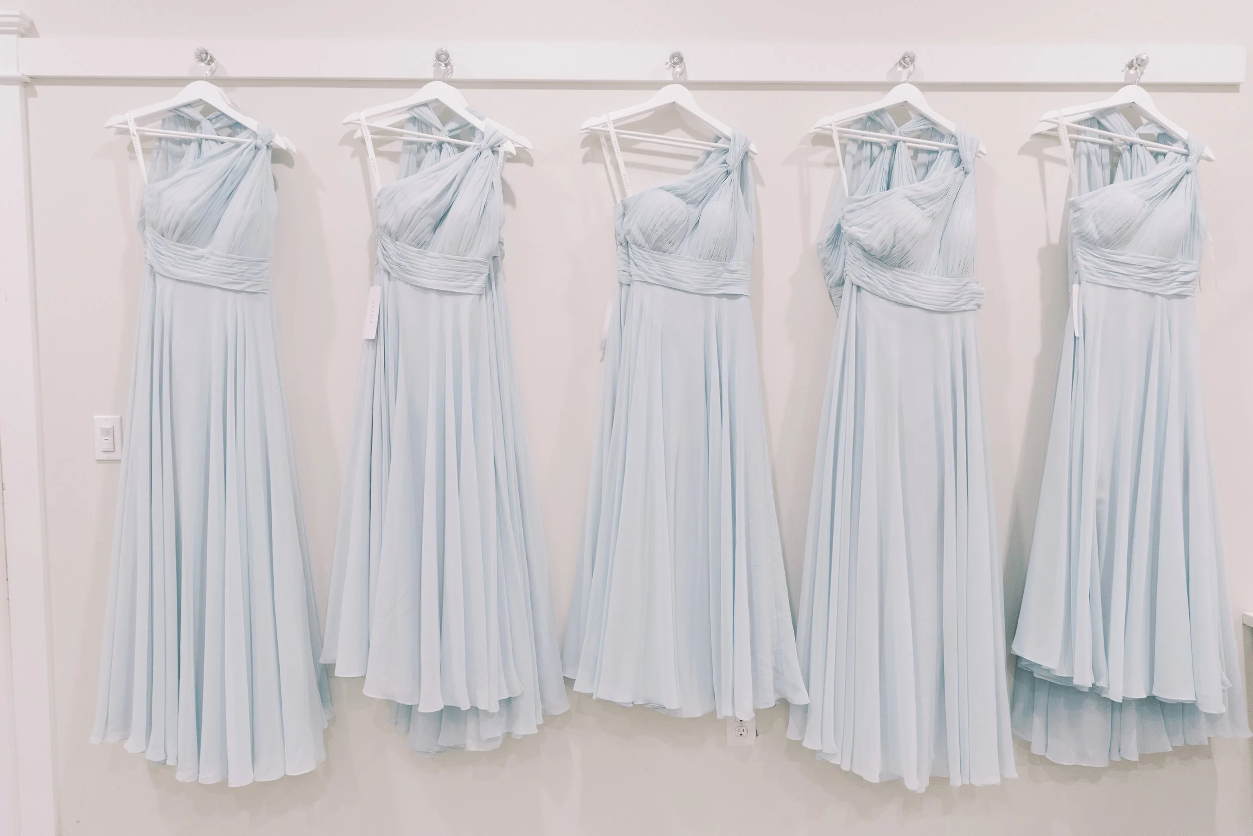 Five identical light blue dresses hanging on white hangers on a white wall.
