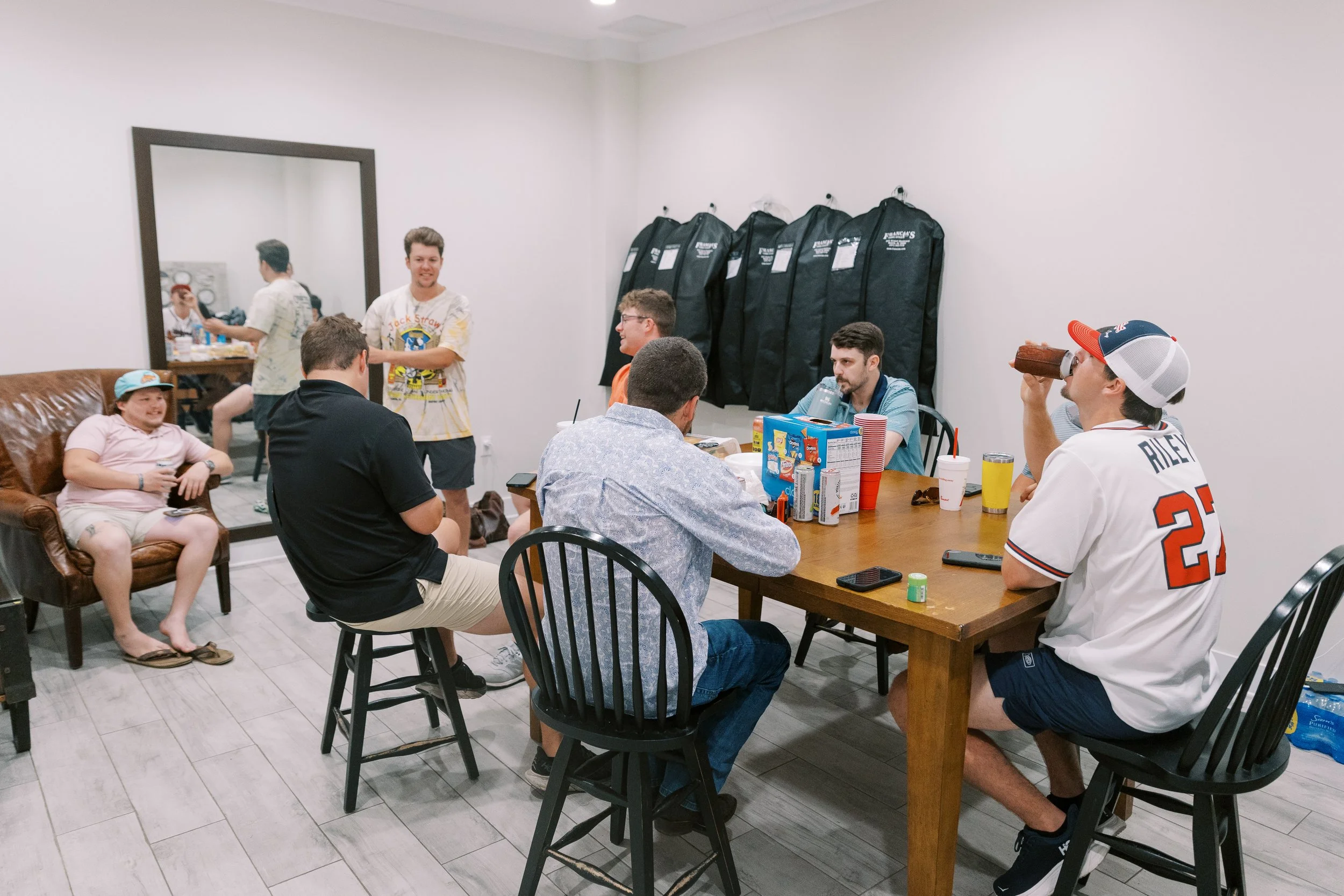 Group of people having a casual gathering in a room with white walls. Several people are seated around a wooden table with snacks and drinks, and others are standing and talking. A young girl is sitting on a brown leather couch wearing a hat. A man i