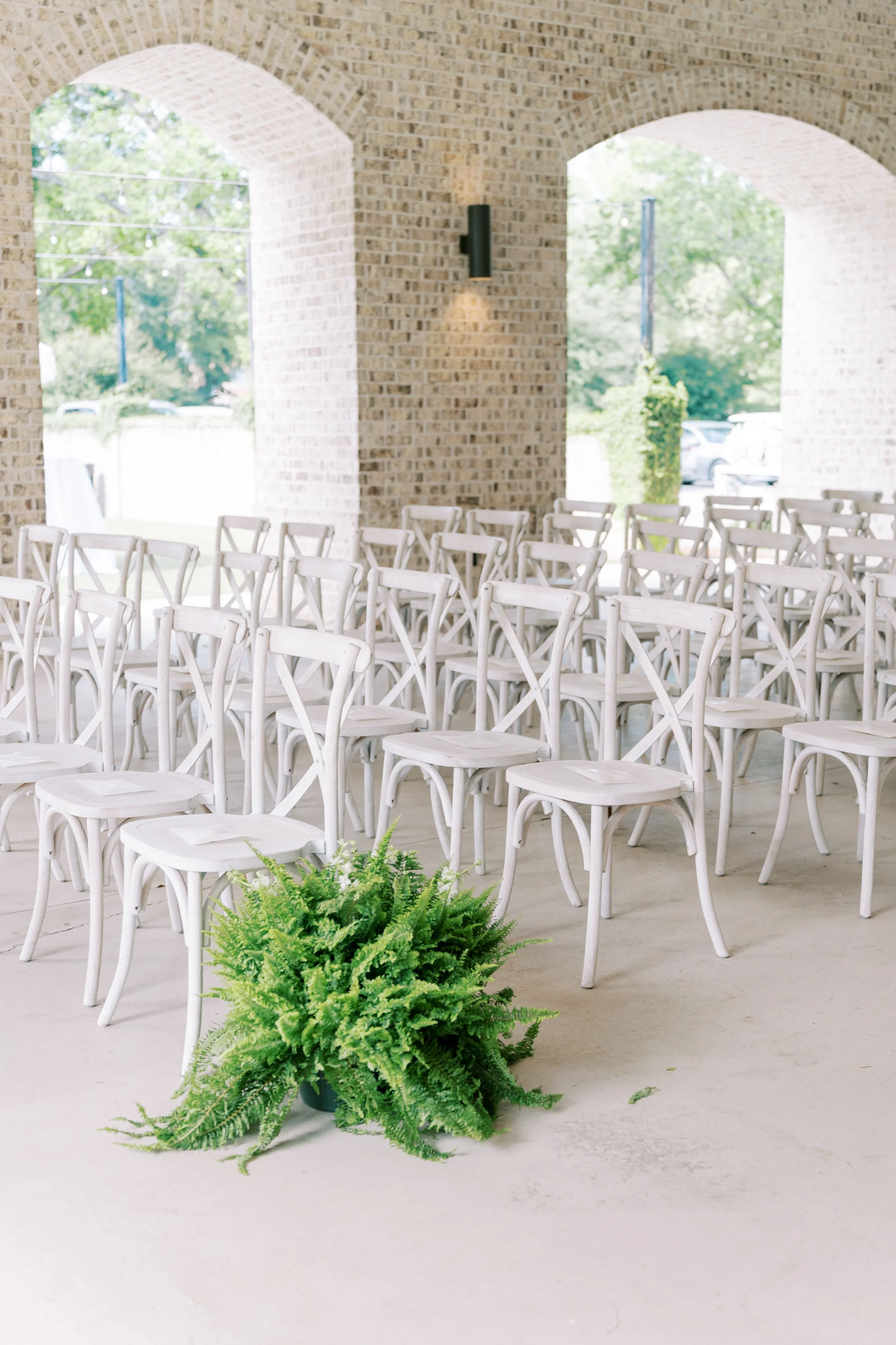 Indoor event space with white chairs arranged in rows and a green fern plant in the foreground, brick walls, arched windows, outdoors visible through the windows.