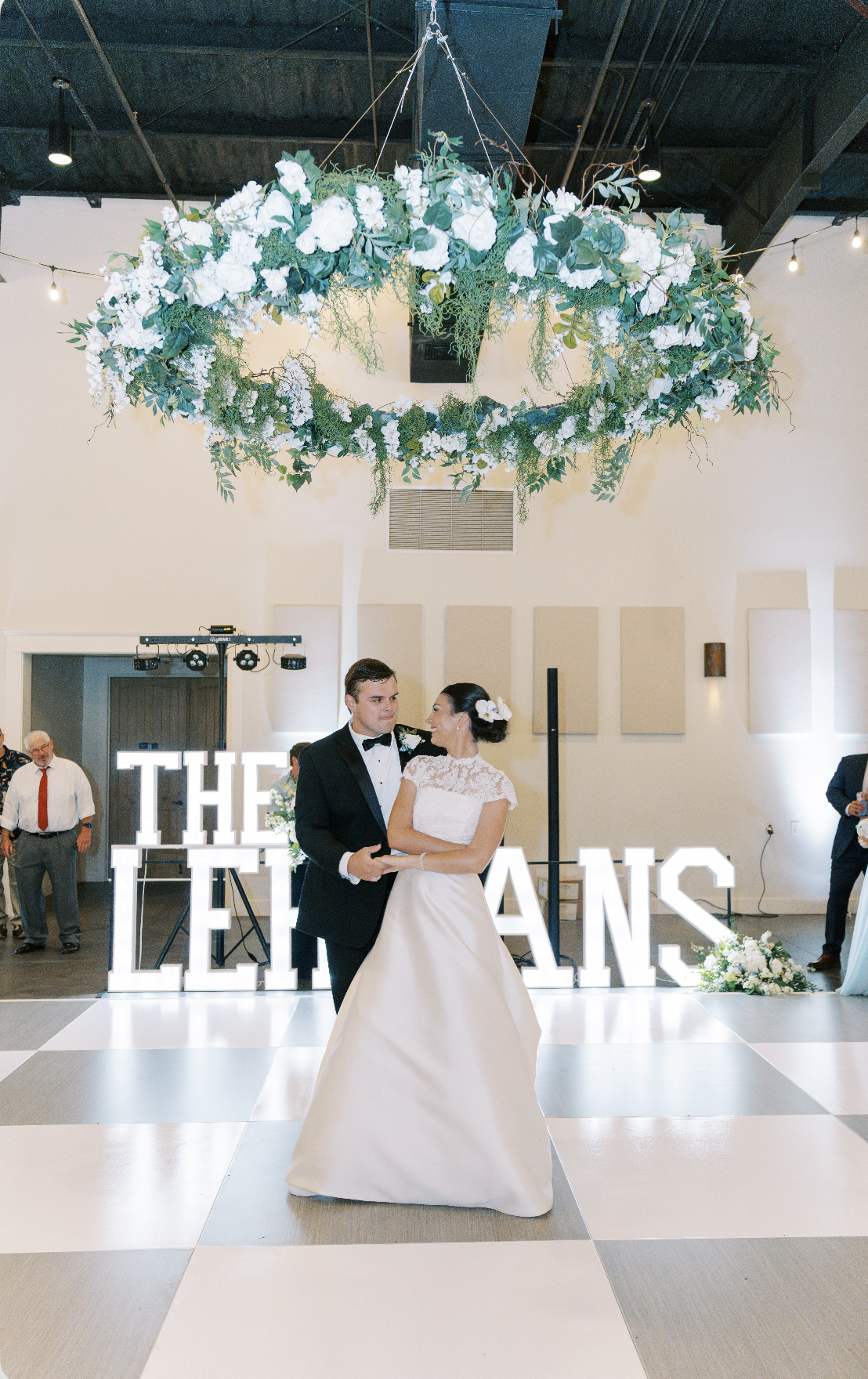 A bride and groom dancing at their wedding reception, with a large illuminated sign reading 'THE LE...' in the background, decorated with a circular floral arrangement hanging from the ceiling.