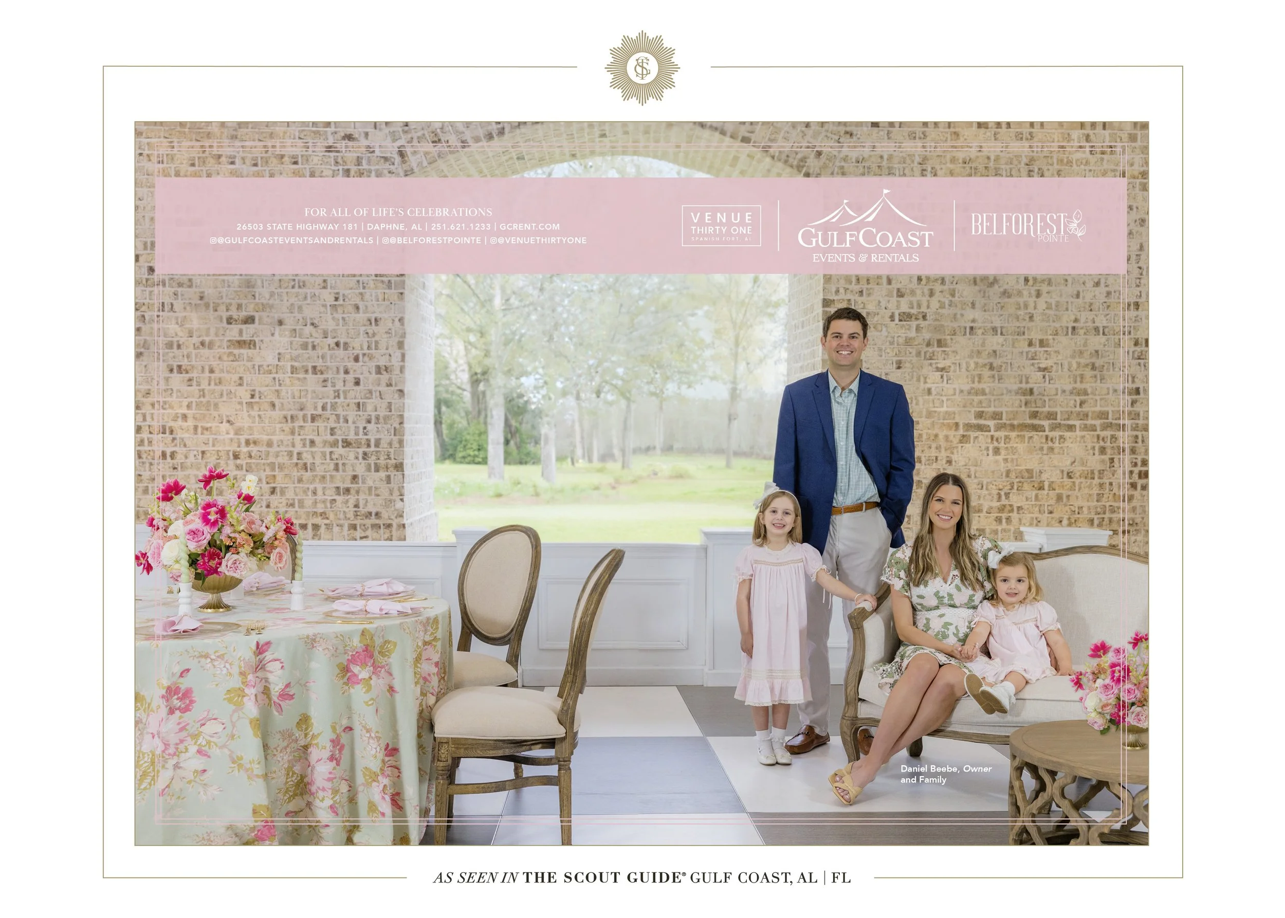 A family of four posing indoors at a celebration venue with brick walls and large windows overlooking a green outdoor area. The family includes a man in a blue blazer, woman in floral dress, and two young girls in pink dresses. There is a table with a floral centerpiece and pink table settings to the left.