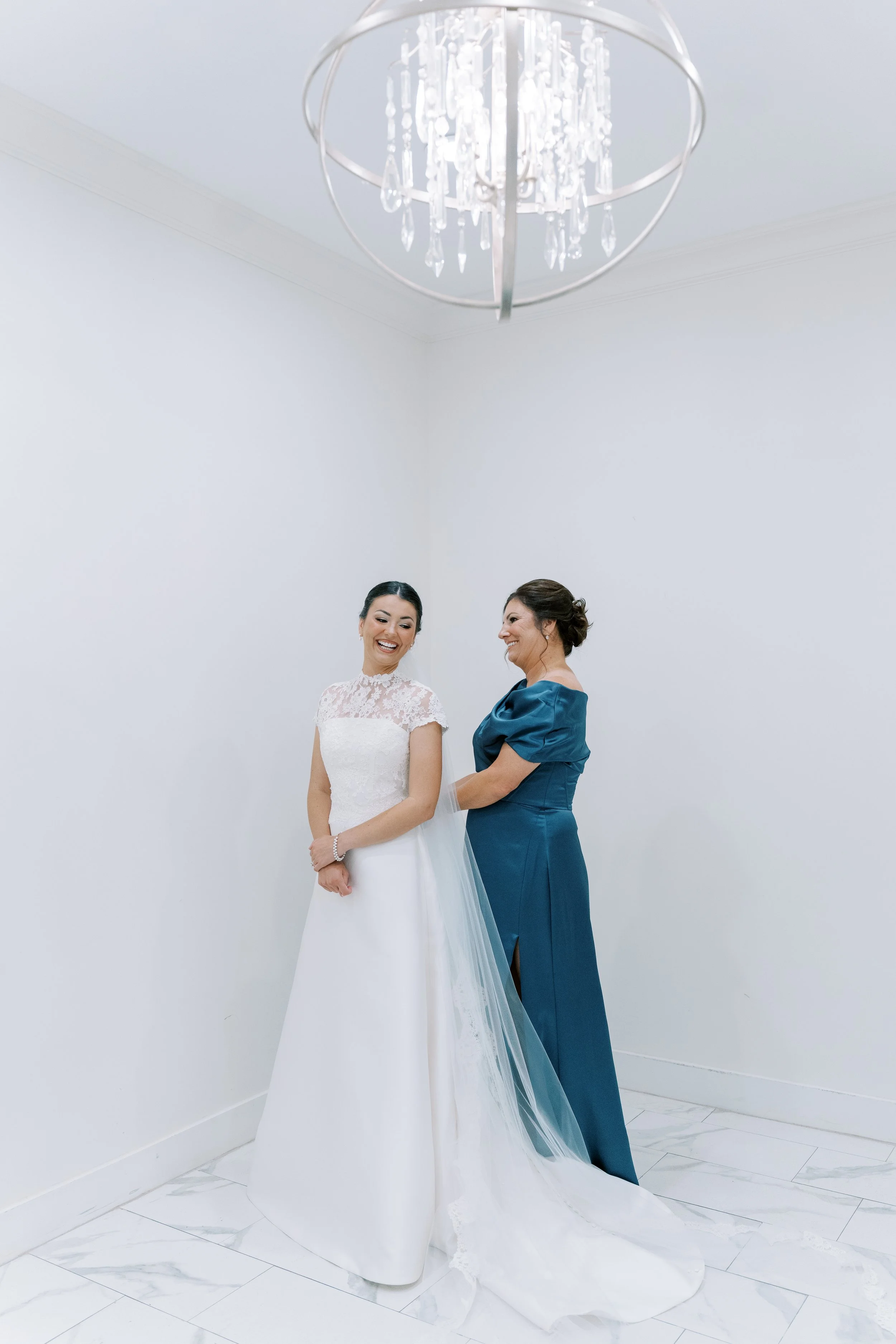 Two women, one in a wedding dress and the other in a teal gown, smiling and holding hands in a minimalistic white room with a chandelier overhead.