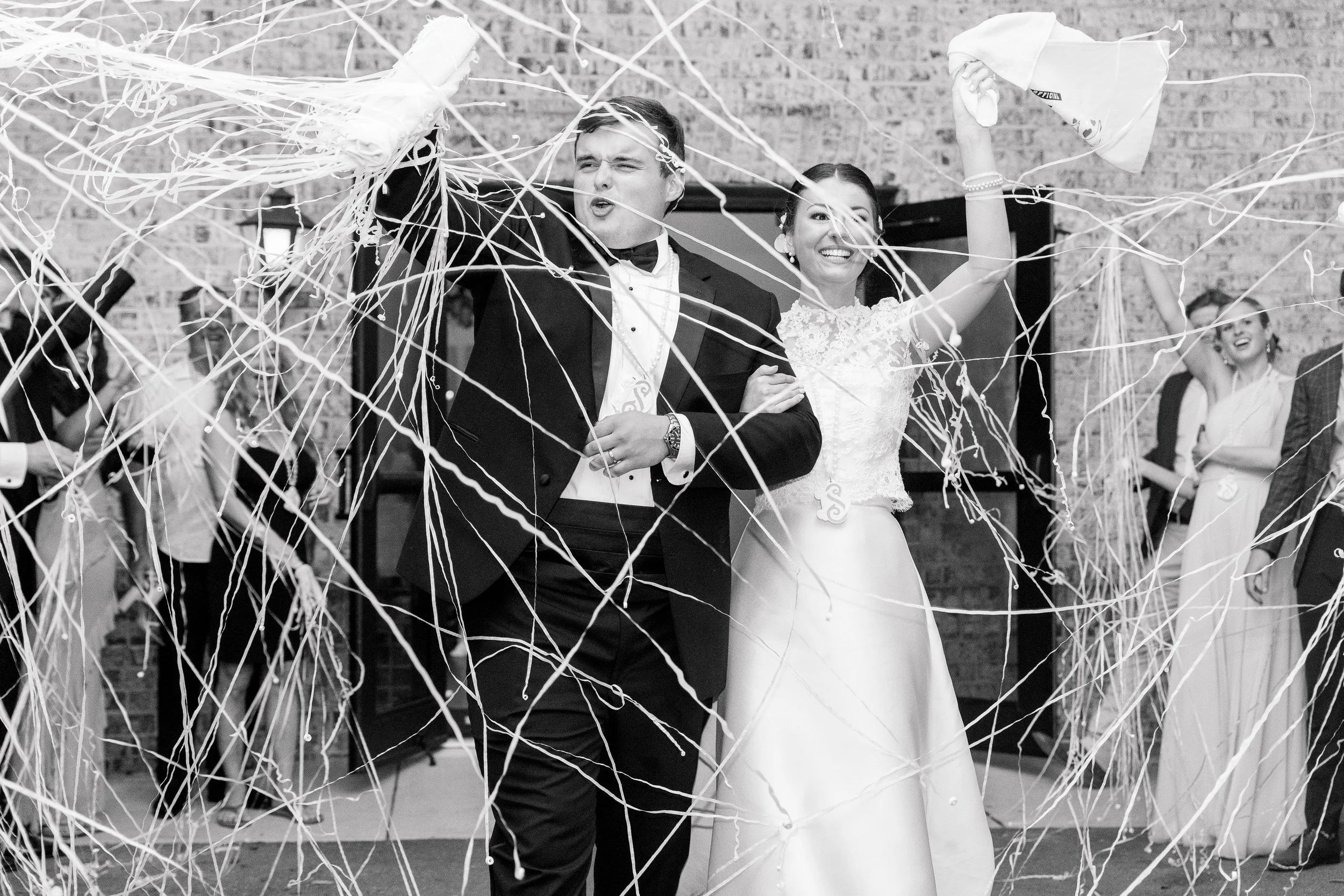 Black and white photo of newlywed couple celebrating with guests, surrounded by streamers at wedding reception.