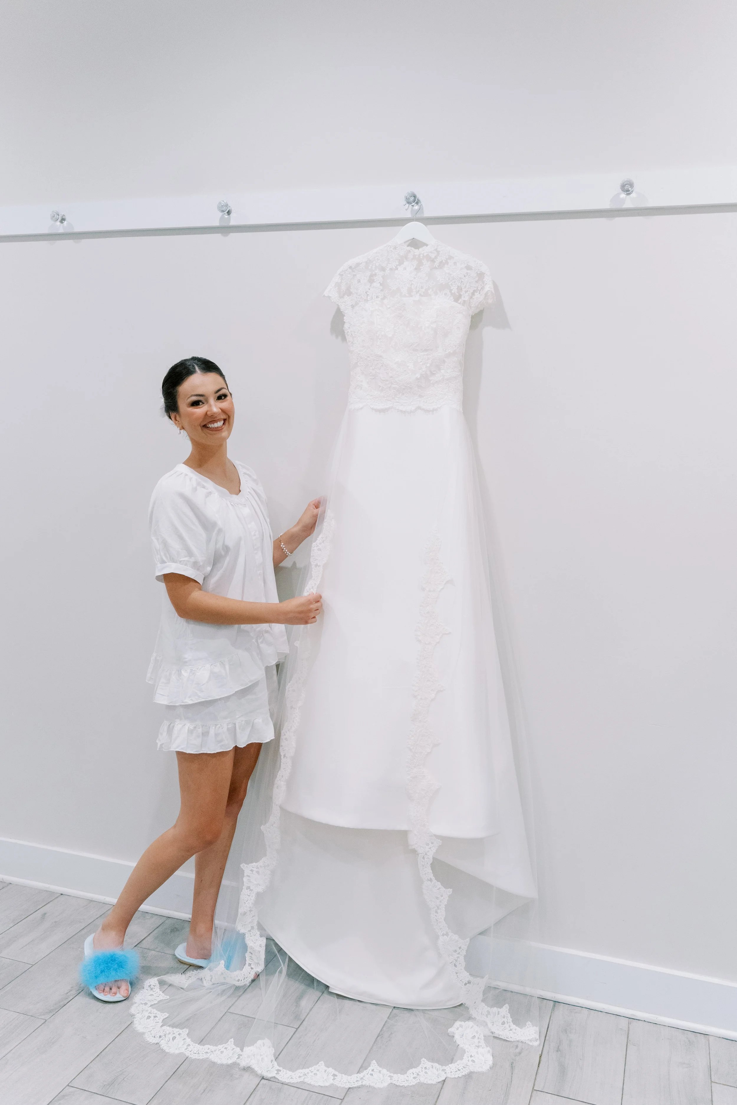 A woman in white pajamas stands next to a white wedding dress hanging on a wall. She is smiling, with her hair tied back, and wearing fluffy slippers.