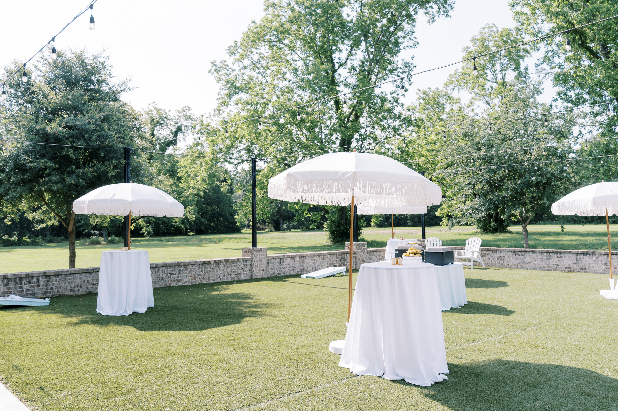 Outdoor event setup with white tables and umbrellas on a grassy lawn, surrounded by trees and string lights.
