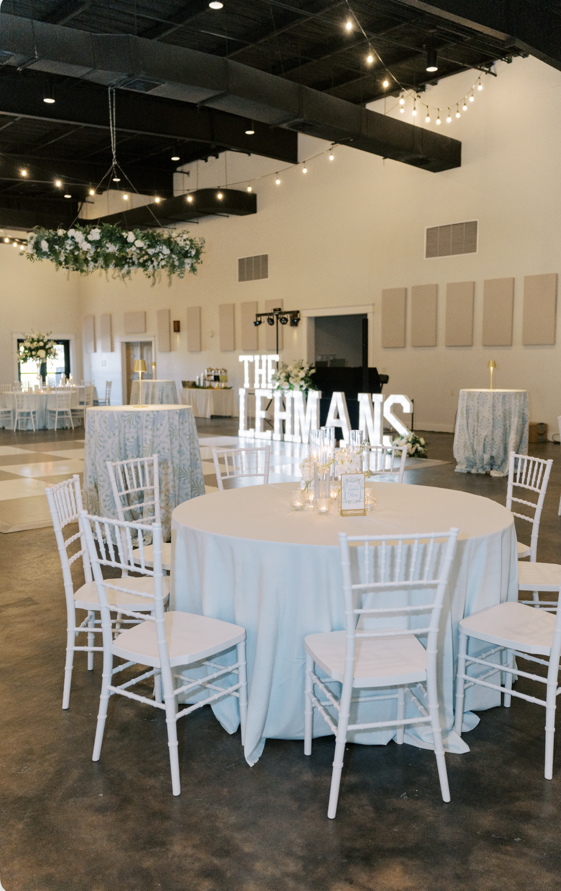 Wedding reception setup with round table covered in white tablecloth, surrounded by white chairs, decorated with candles and small flower arrangements. Large illuminated sign displaying "THE LEHMANS" is in the background. Decor includes hanging strin