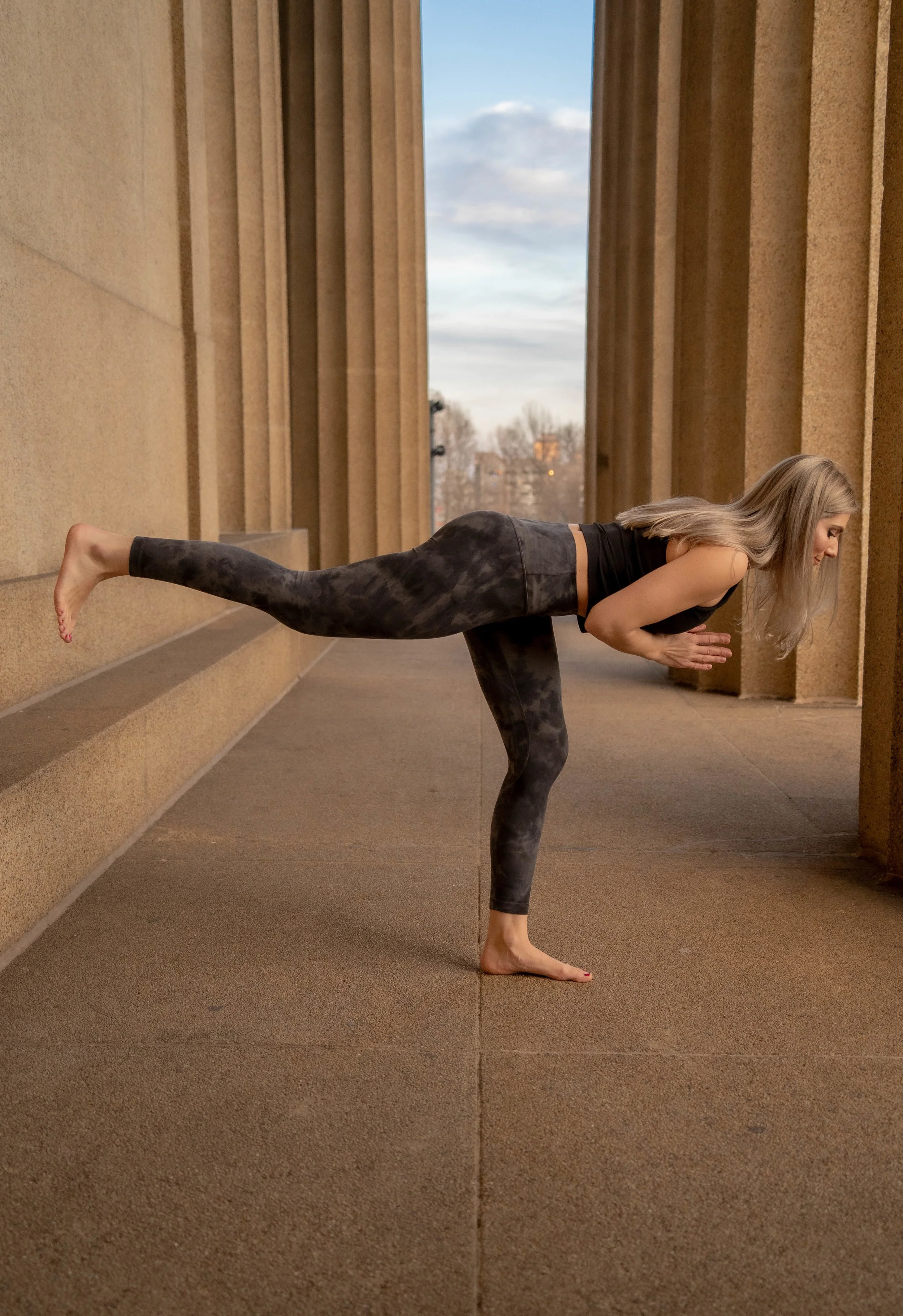 A woman practicing yoga outdoors, balancing on one leg with her other leg extended behind her, leaning forward with her hands together in prayer position.