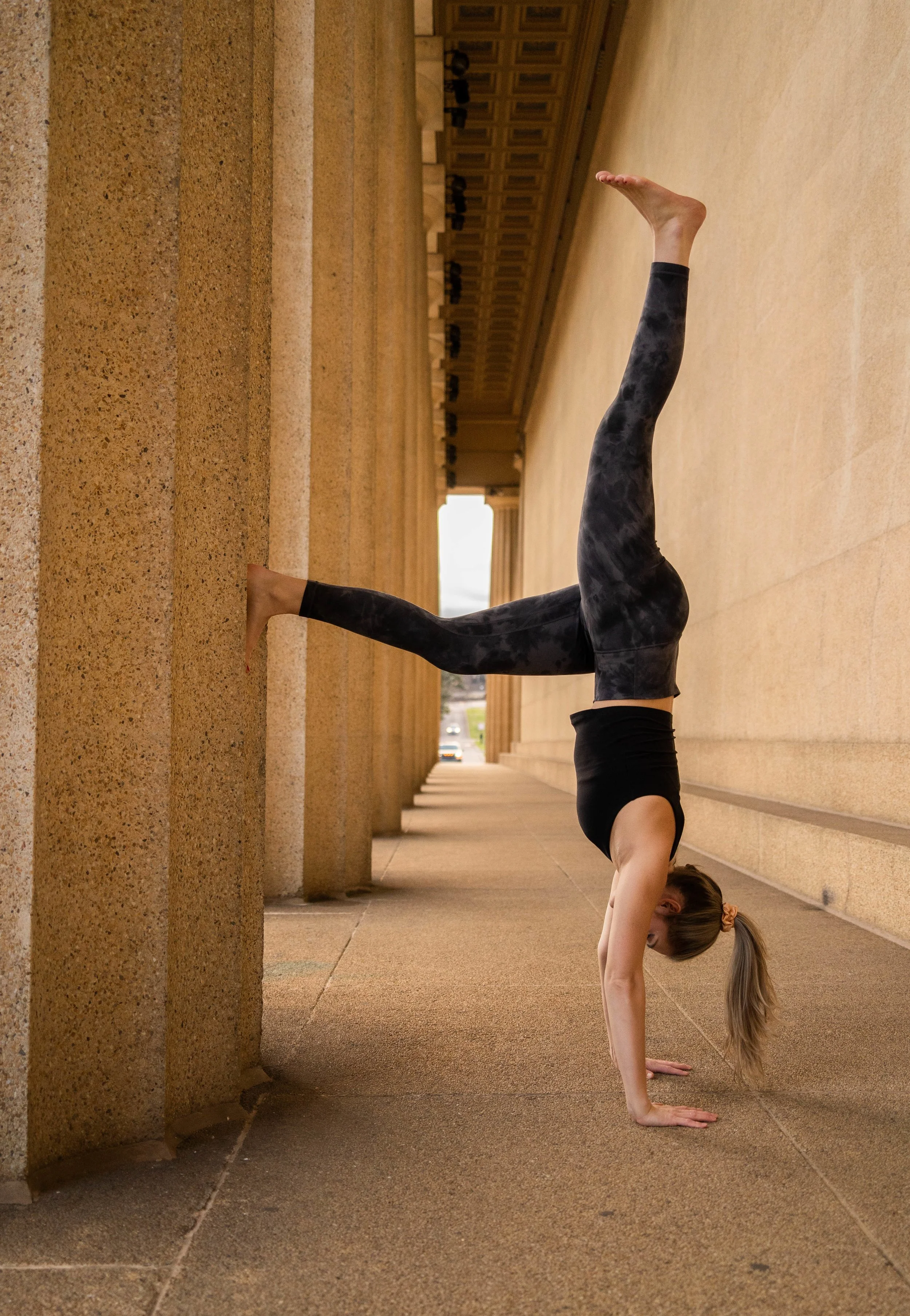 A woman in athletic wear performing a handstand against a stone wall in an outdoor architectural setting with columns.