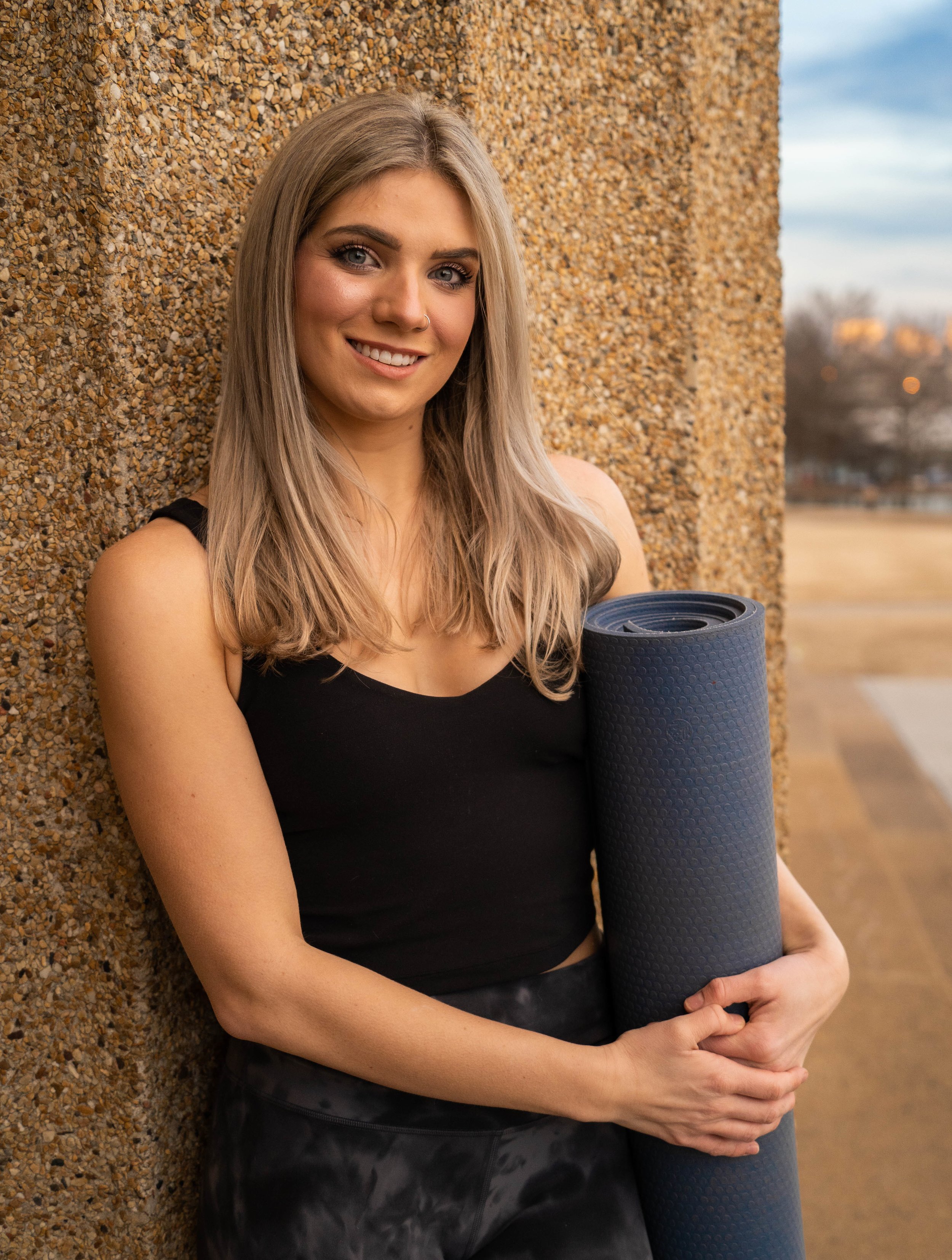 A young woman with blonde hair, blue eyes, and a nose ring, smiling, holding a rolled-up yoga mat, leaning against a textured brown wall outdoors.