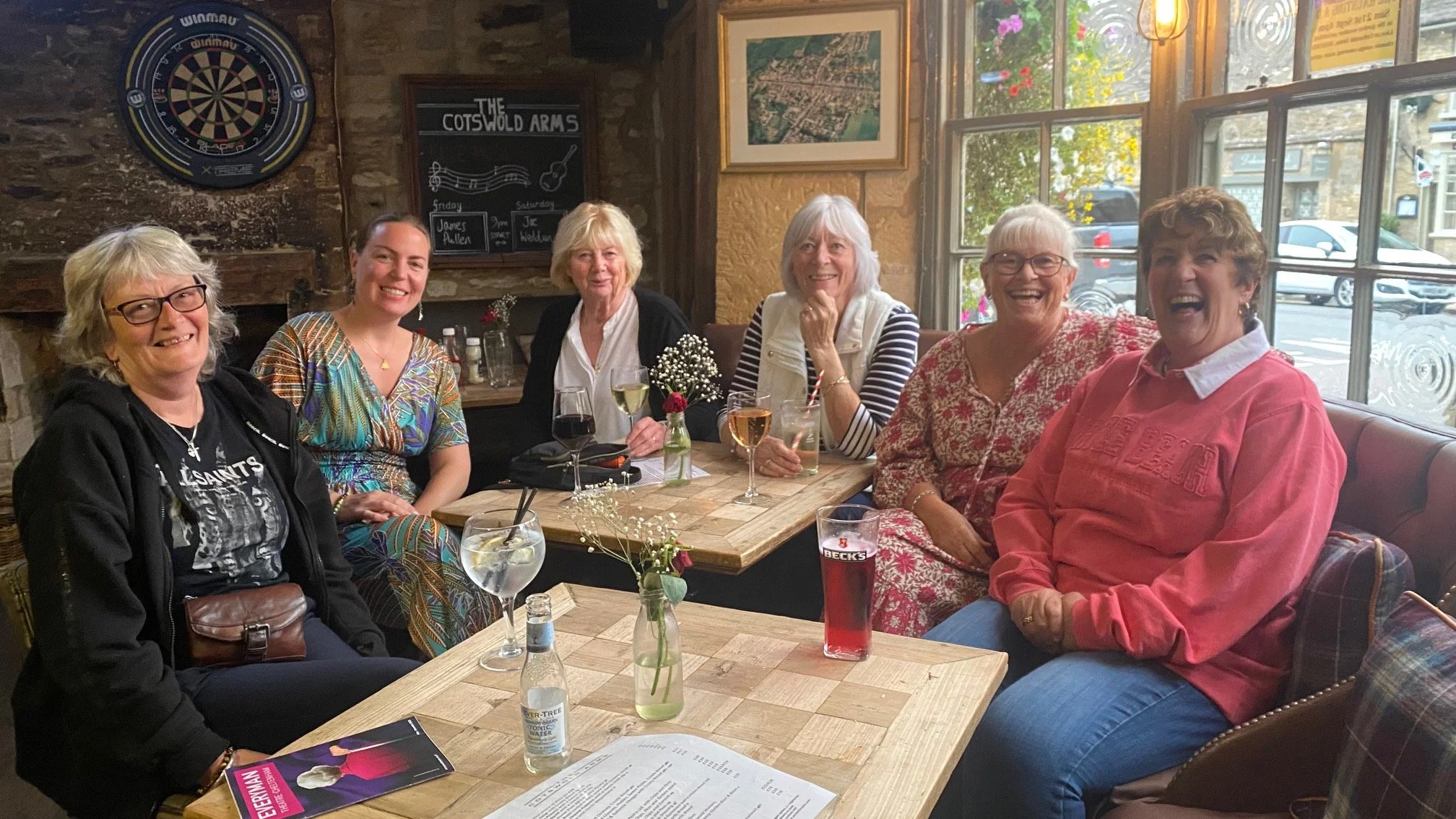 Seven women sitting at a wooden table in a cozy restaurant, smiling and laughing. The table has various drinks, a small floral centerpiece, and menus. The background features a dartboard, a chalkboard, and a large window with a view of parked cars and a sidewalk outside.