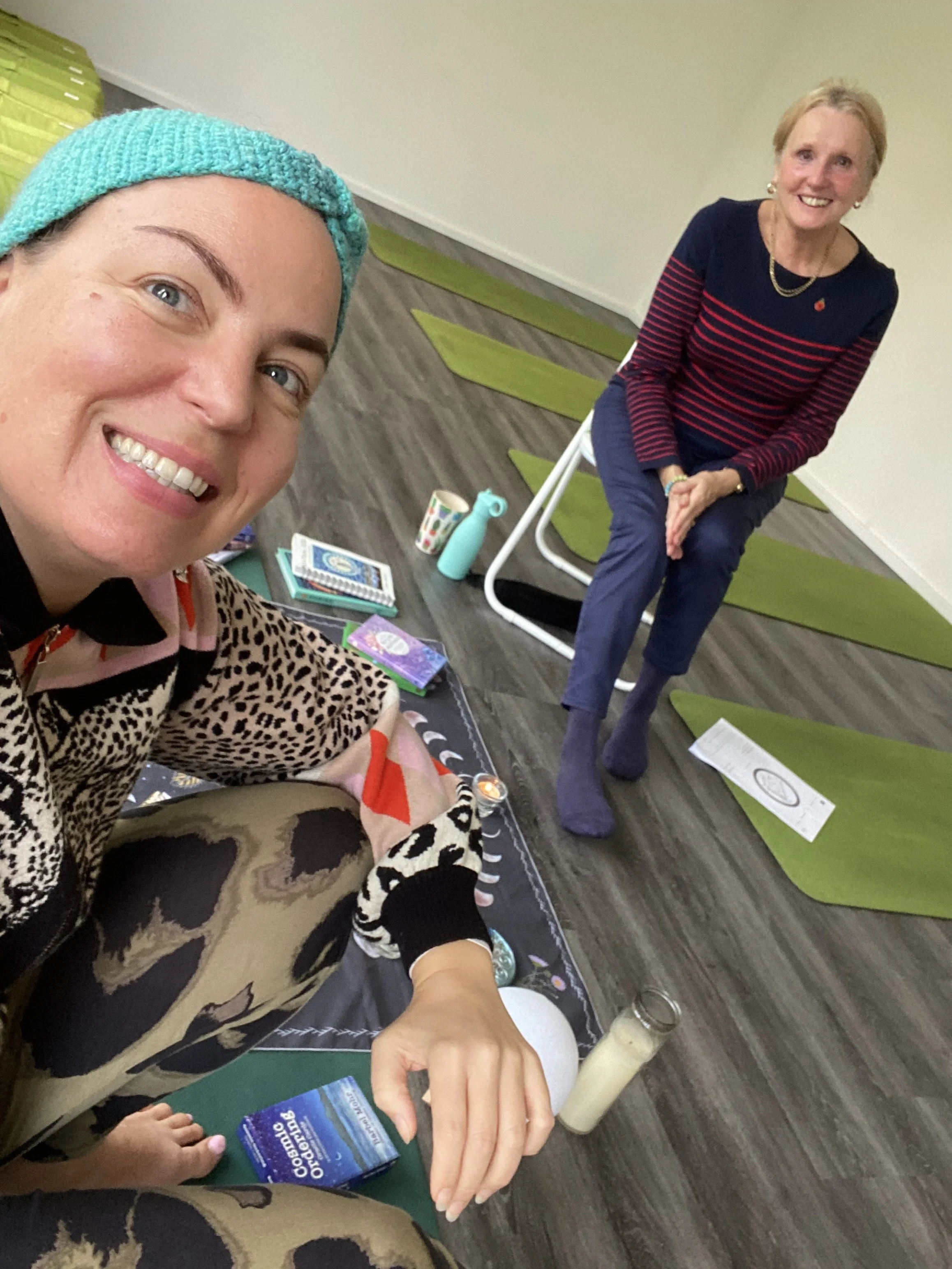 Two women at a wellness or yoga class with yoga mats, candles, and wellness items, smiling and taking a selfie.