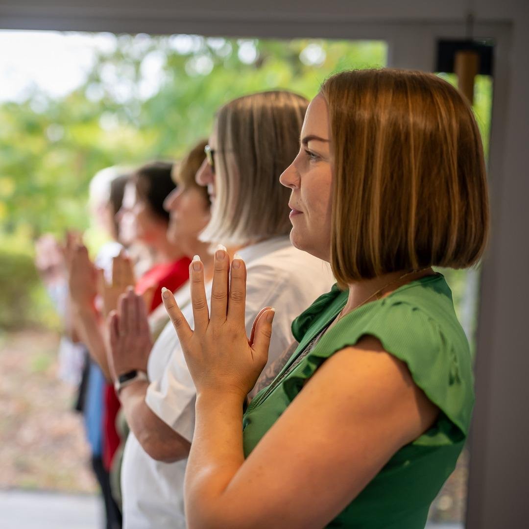 A group of women standing in a line indoors, praying with their eyes closed and hands raised, with a window showing green trees outside.