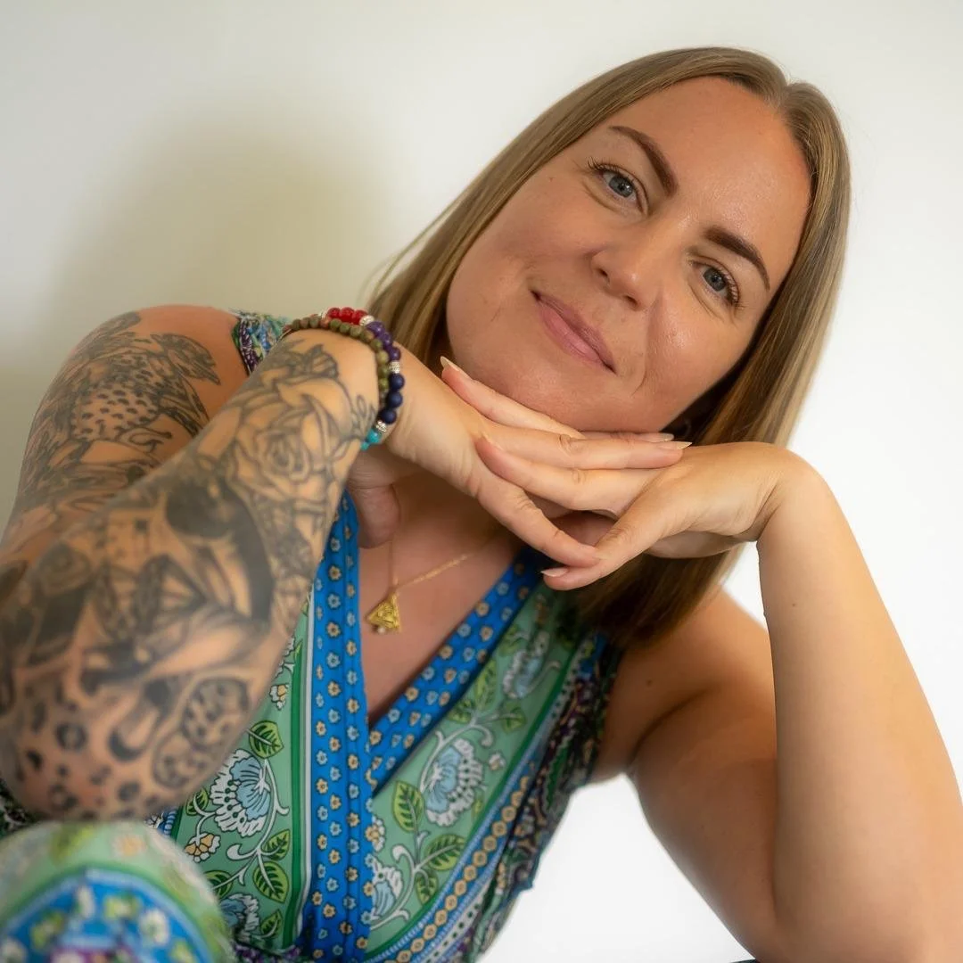 A woman with long brown hair and blue eyes, smiling, resting her chin on her hands. She has colorful tattoos on her arms and is wearing a sleeveless blouse with a floral and geometric pattern. She also has beaded bracelets on her wrist and a gold necklace with a triangular pendant.