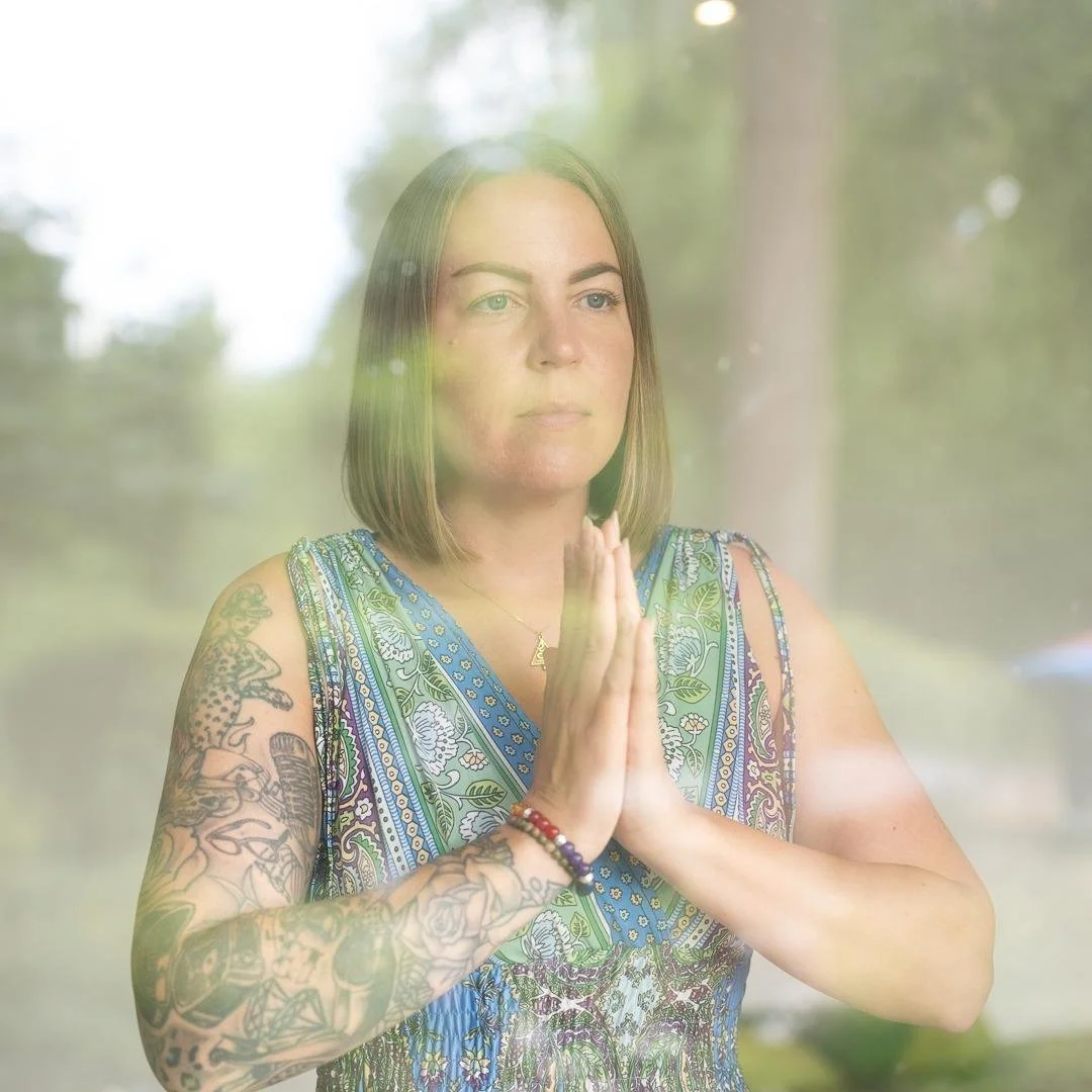 A woman with shoulder-length brown hair and tattoos on her left arm wearing a sleeveless colorful patterned top, standing outside with her hands pressed together in a prayer position, seen through a glass window.