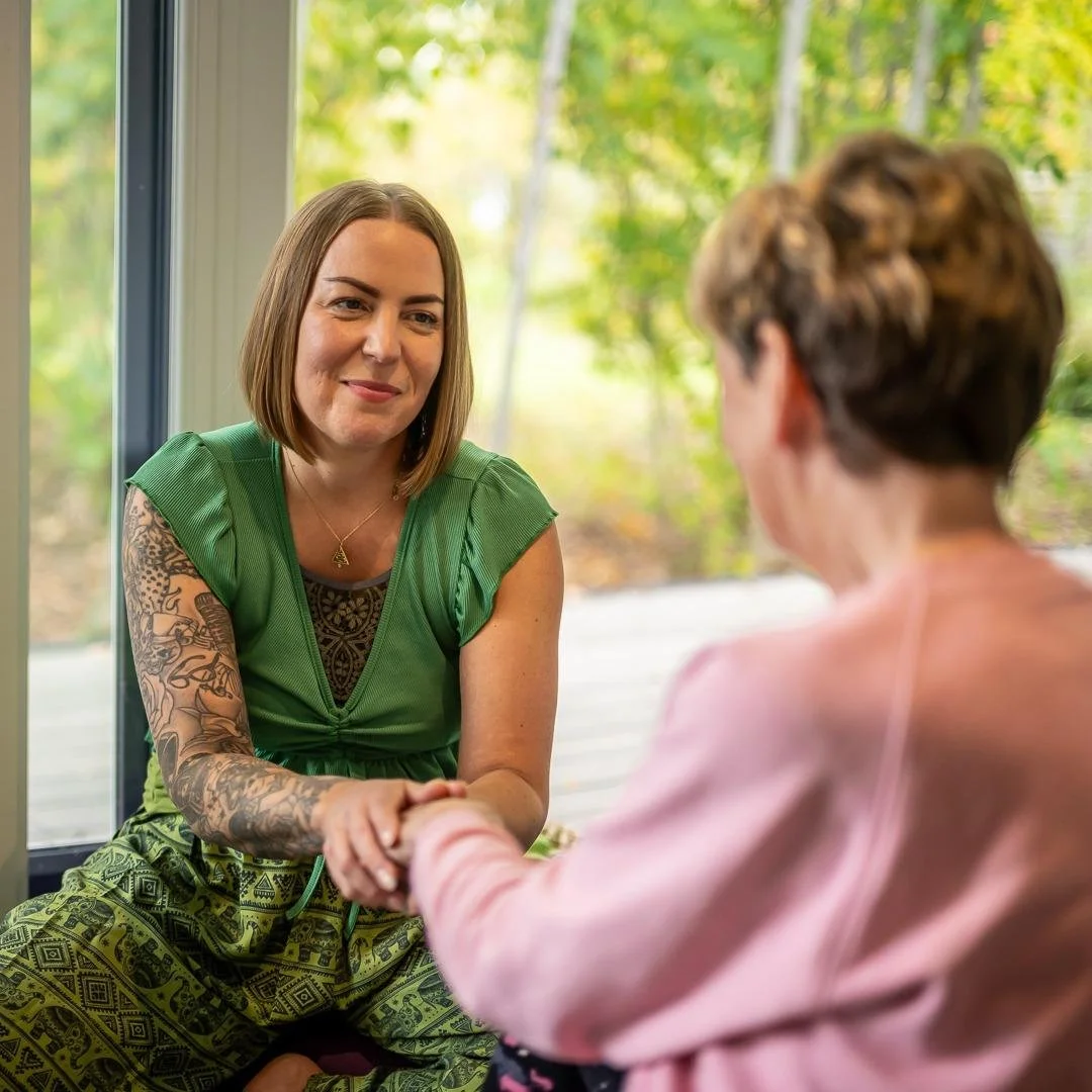 Two women having a conversation indoors, with one woman smiling and holding the other's hand. The woman facing the camera has shoulder-length brown hair, tattoos on her arm, and is wearing a green top with patterned green pants. The woman with her back to the camera has short curly hair and is wearing a light pink blazer. They are near a large window with greenery outside.