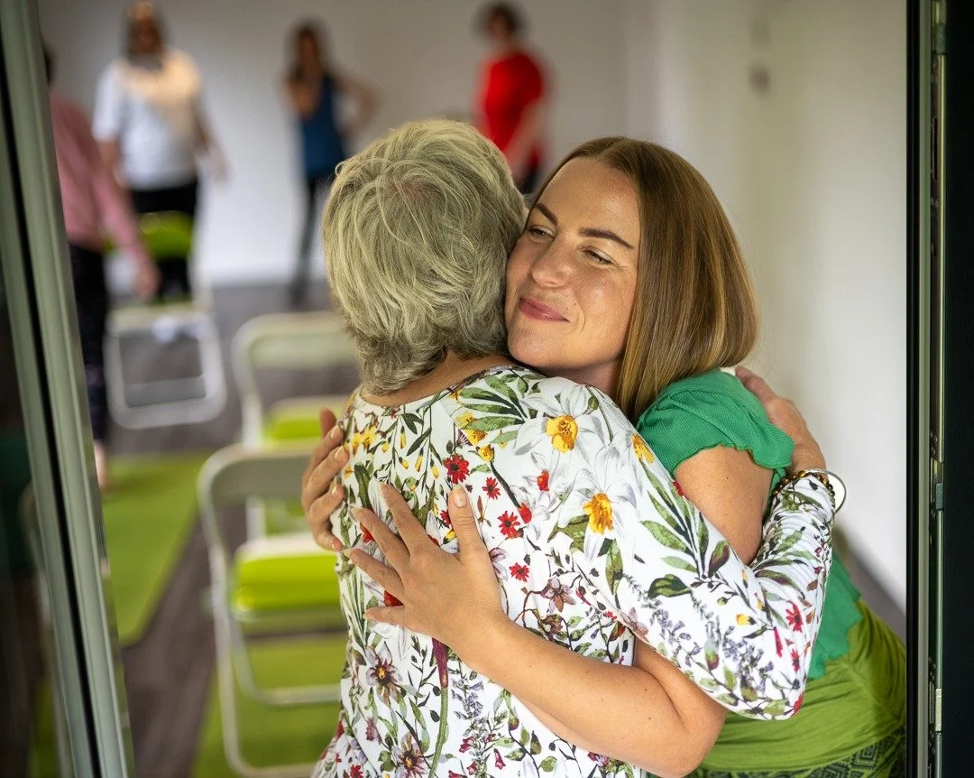 Two women hugging, the younger woman with long brown hair wearing a green top, the older woman with short gray hair wearing a floral blouse, in a room with chairs and blurred people in the background.