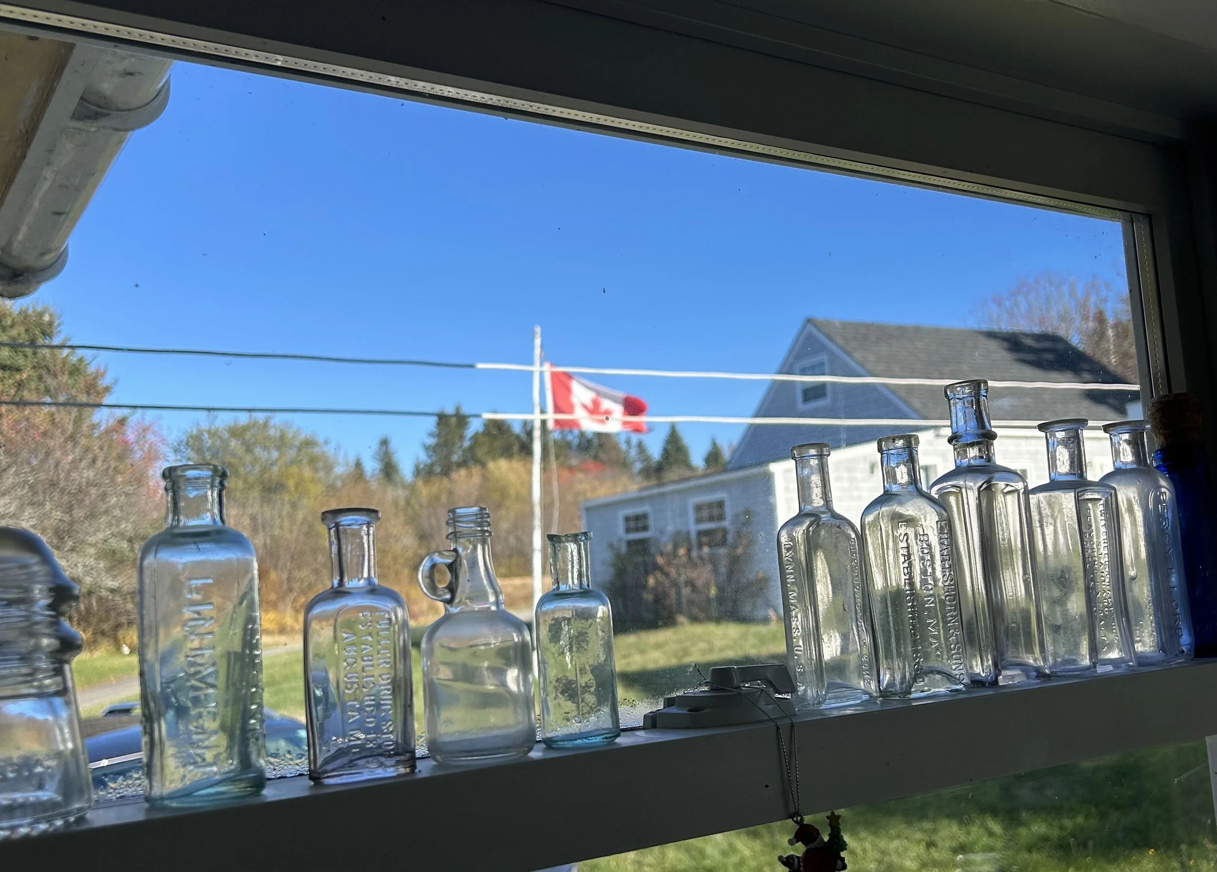 View from a window with glass bottles lined on the window sill; a Canadian flag flying outside against a blue sky, with a house and trees in the background.