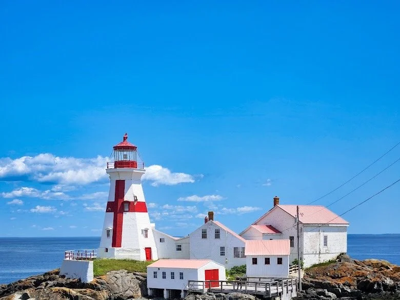 Lighthouse on rocky coastline with white buildings, red roofs, and a blue sky with scattered clouds.