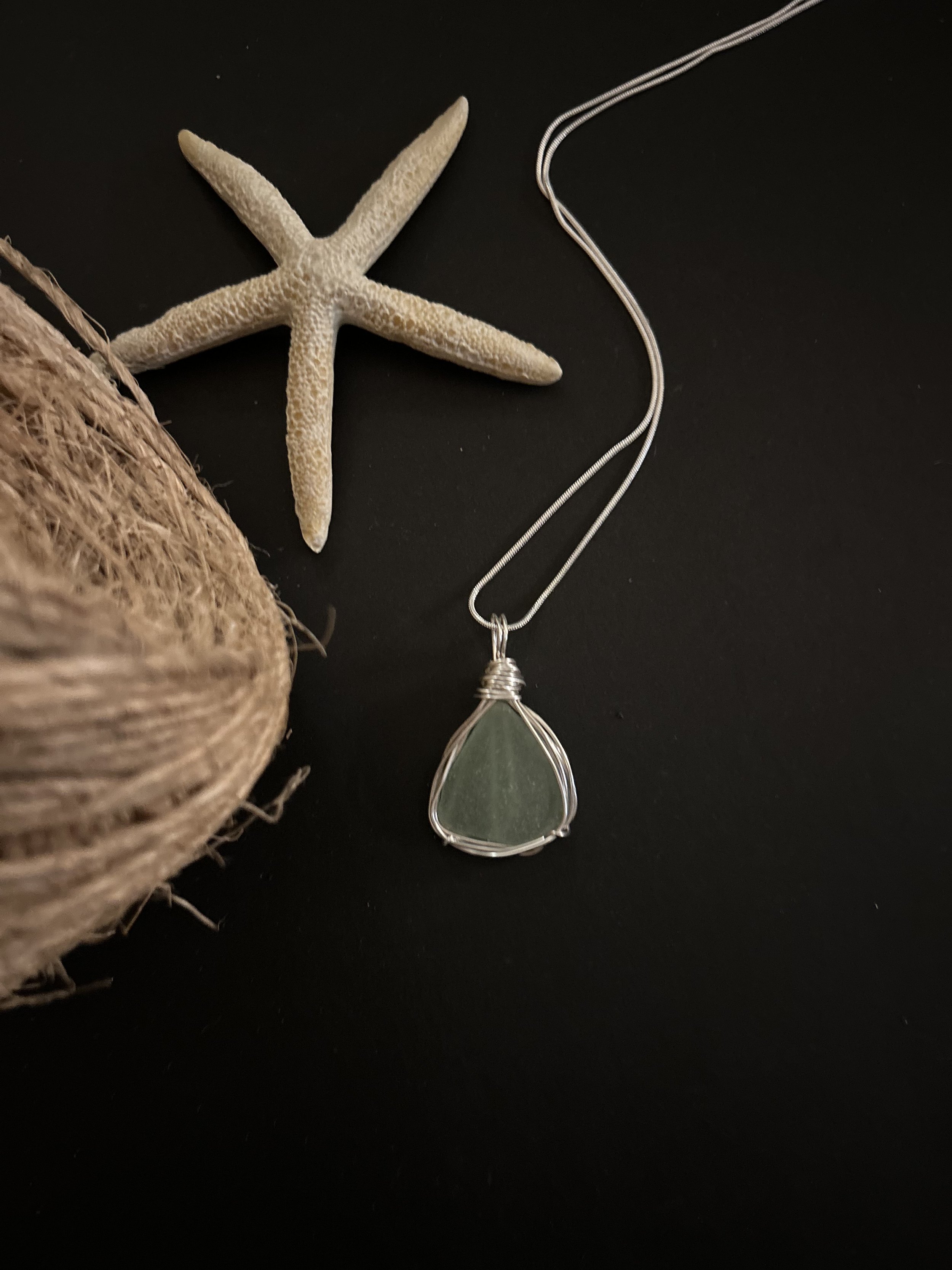 Sea glass pendant necklace with silver wire wrapping, starfish, and a ball of twine on a black background.