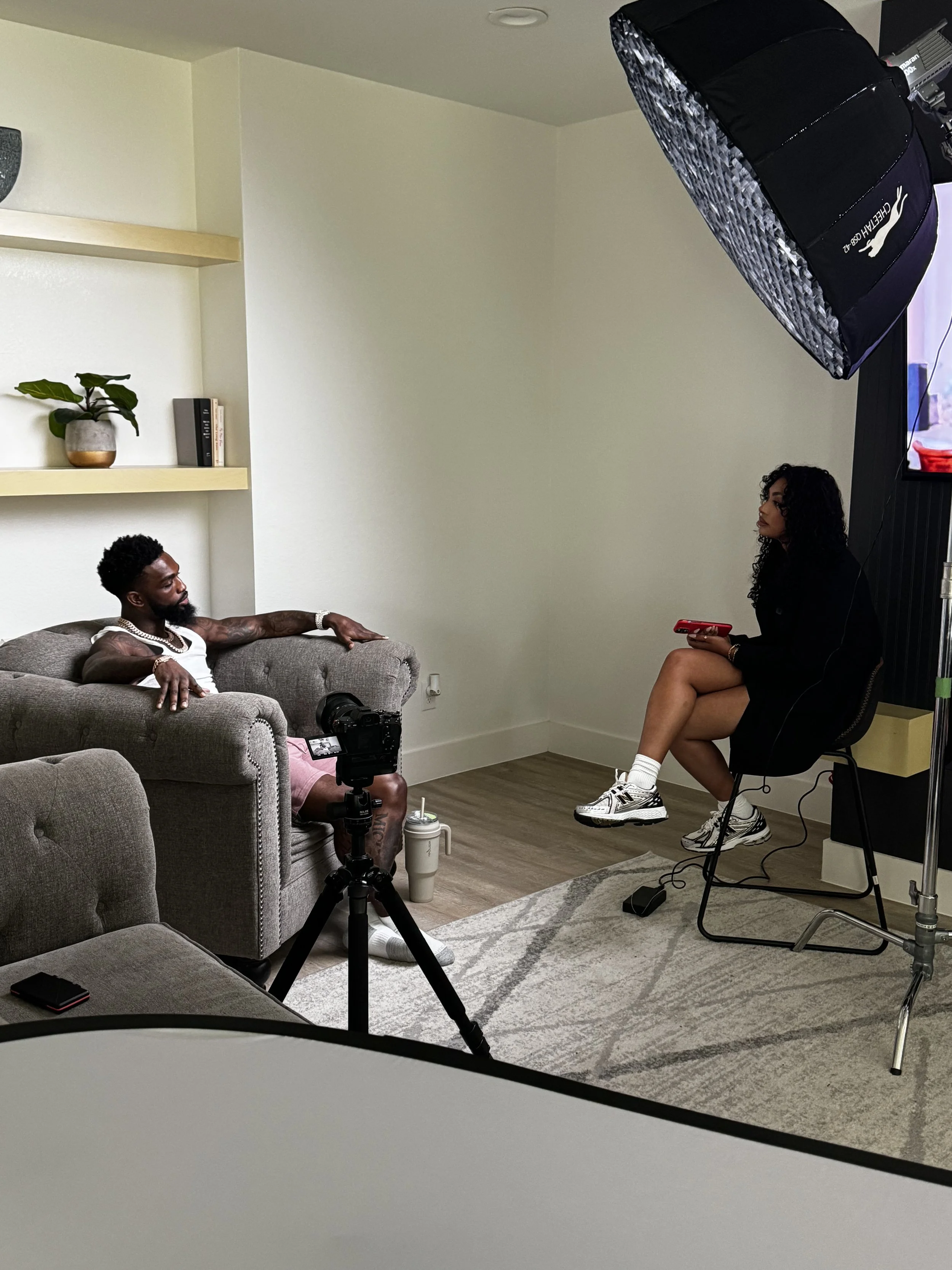 A woman sitting on a chair with a notebook, being interviewed by a man on a sofa, with professional lighting and camera equipment for a video shoot in a room with a bookshelf and houseplant.