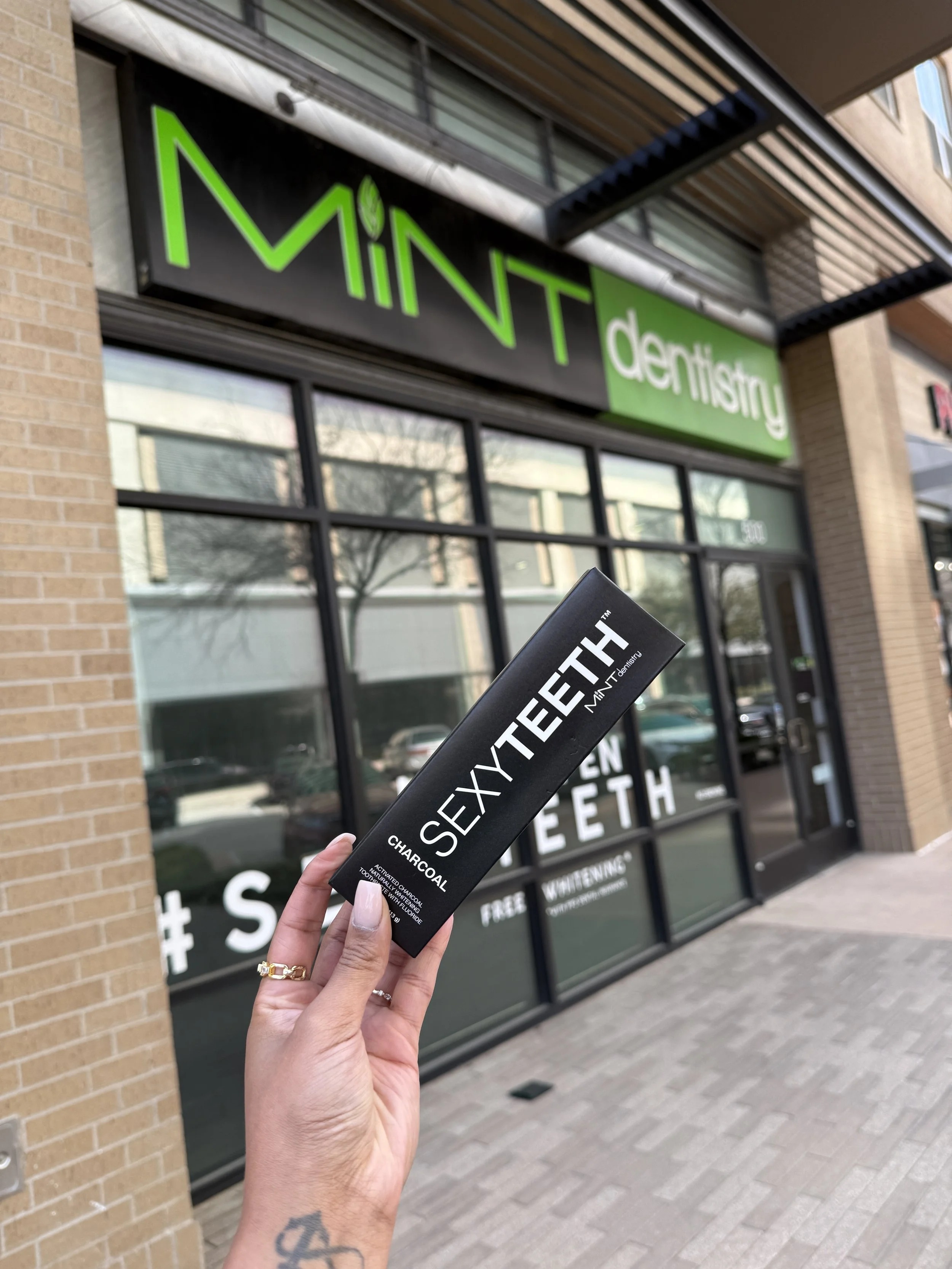 Person holding a black box of SEXYTEETH charcoal activated dental floss in front of a dental office with a green and black sign that reads 'MINT dentistry'.