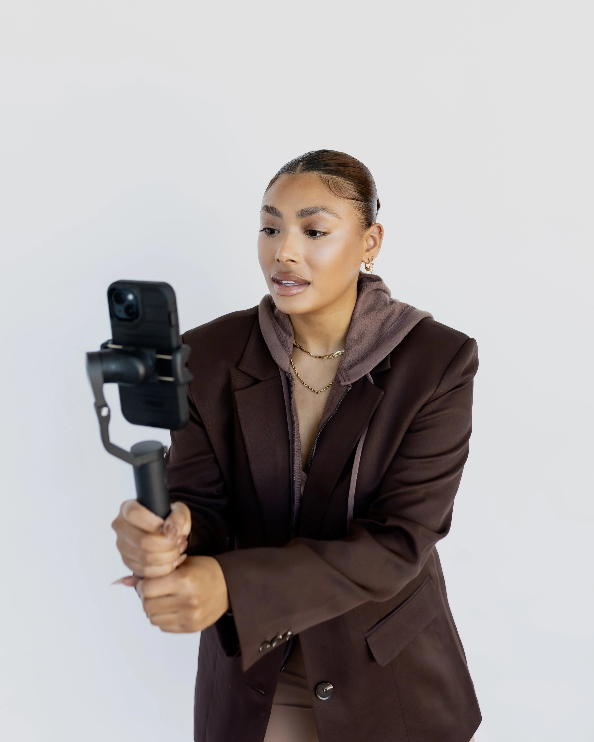 A young woman with brown hair in a bun wearing a brown blazer, gold jewelry, and a brown hoodie, holding a camera stabilizer with a smartphone, capturing a video or taking a selfie against a white background.