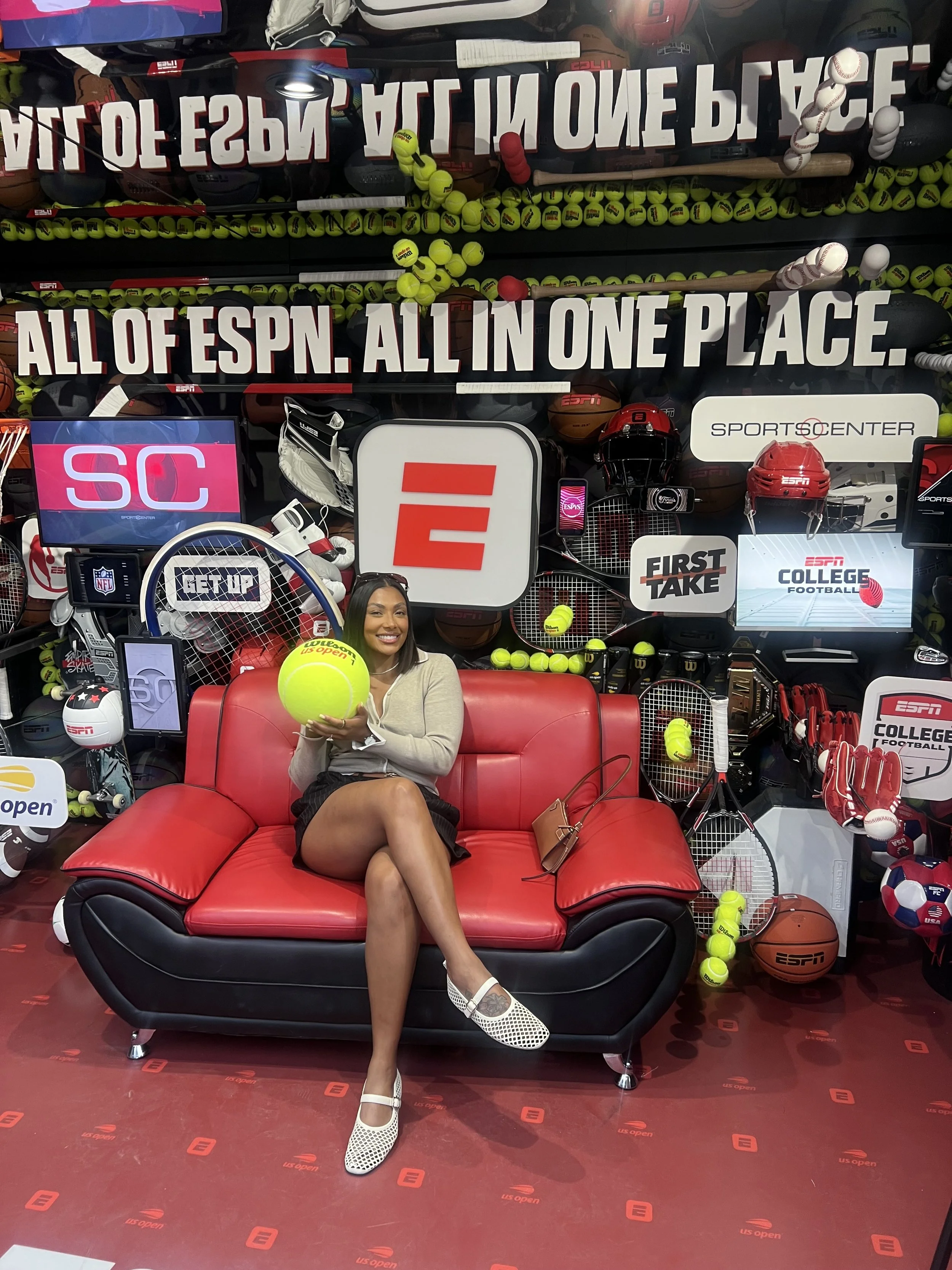 A woman sitting on a red and black couch, smiling and holding a large tennis ball. Behind her is a display with sports equipment, tennis balls, and ESPN logos, with a backdrop of tennis balls.