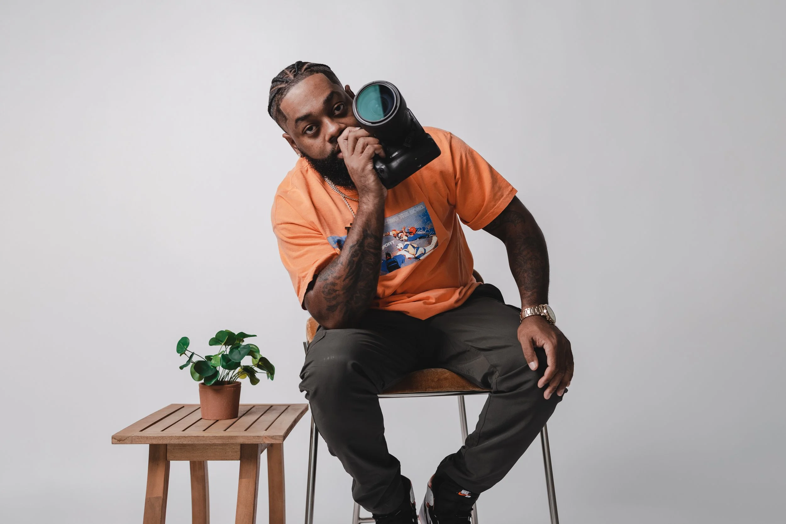 A man sitting on a chair holding a camera to his eye, wearing an orange t-shirt, black pants, and a watch, with a small potted plant on a wooden table beside him, against a plain gray background.
