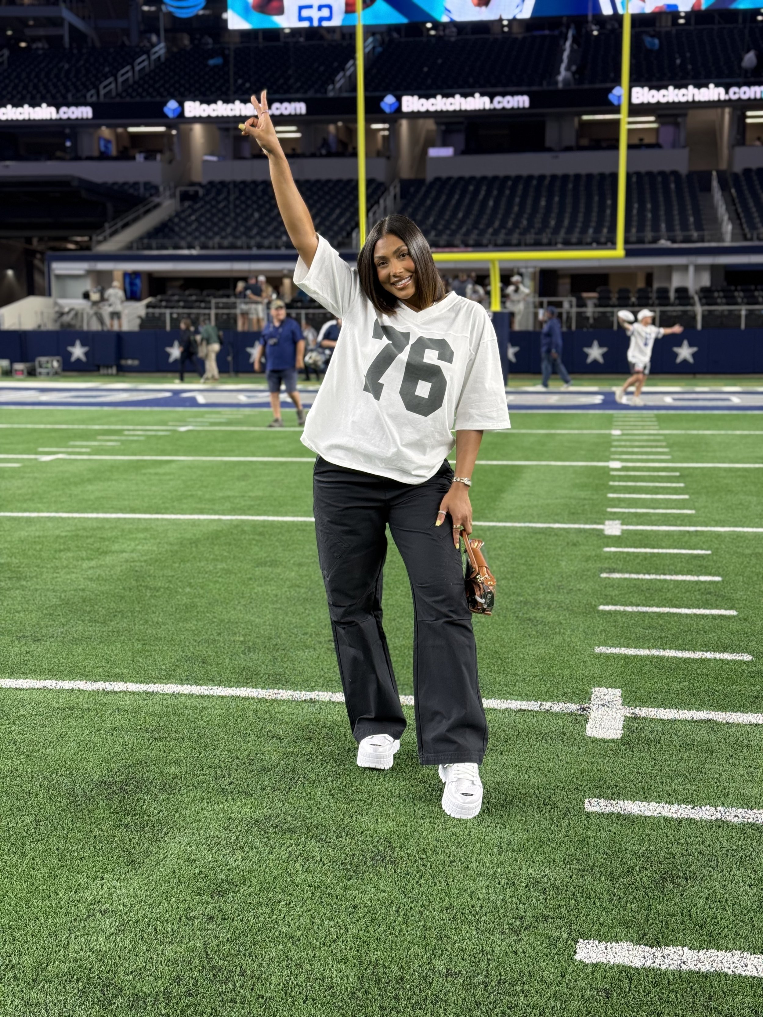 A smiling woman on a football field, wearing a white jersey with the number 76, holding sunglasses, and making a peace sign with her right hand. The stadium seats and scoreboard are visible in the background.
