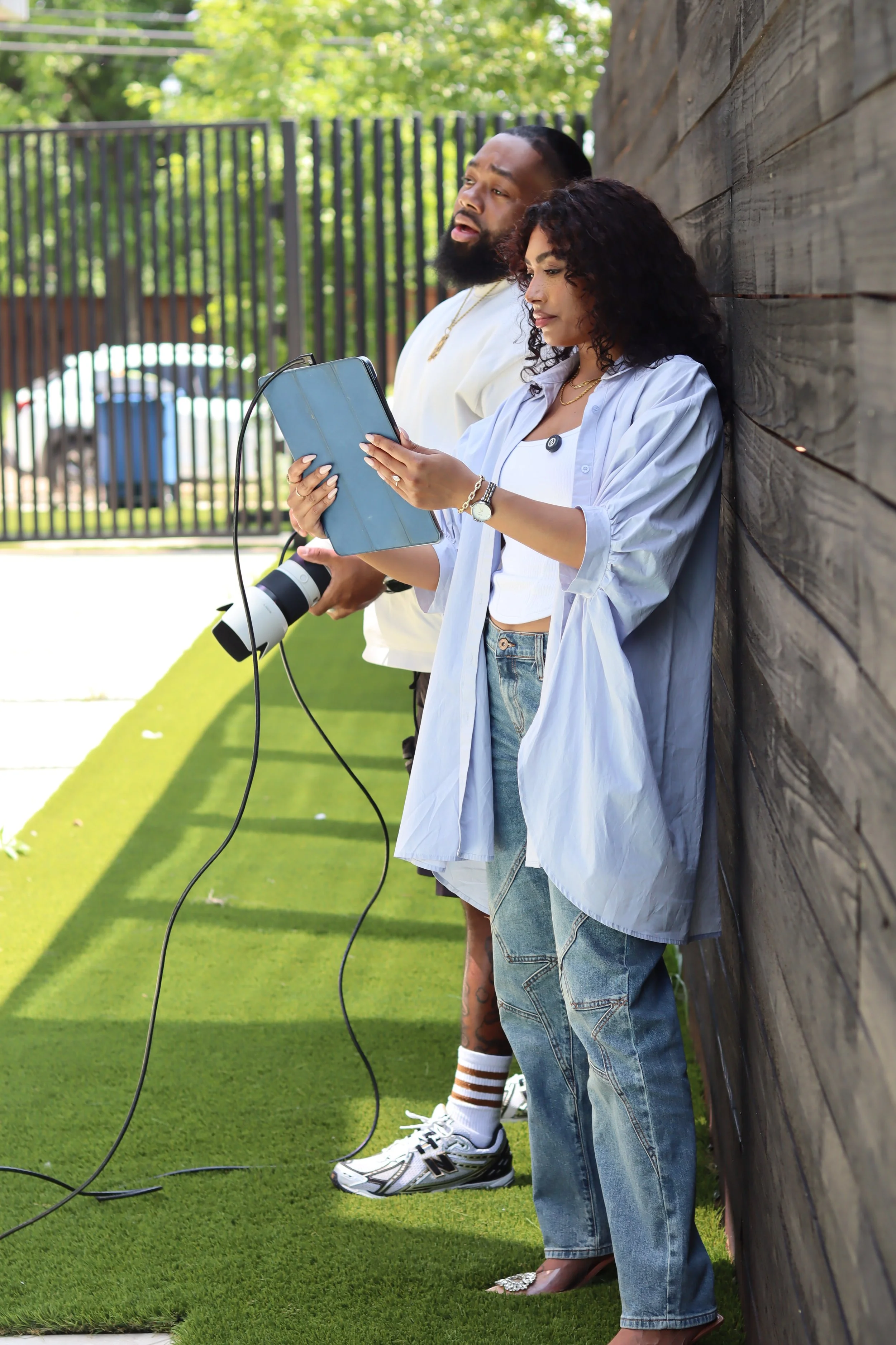 Two people standing outdoors near a wooden wall, one woman using a tablet, and one man holding a camera, with a black metal fence and trees in the background.