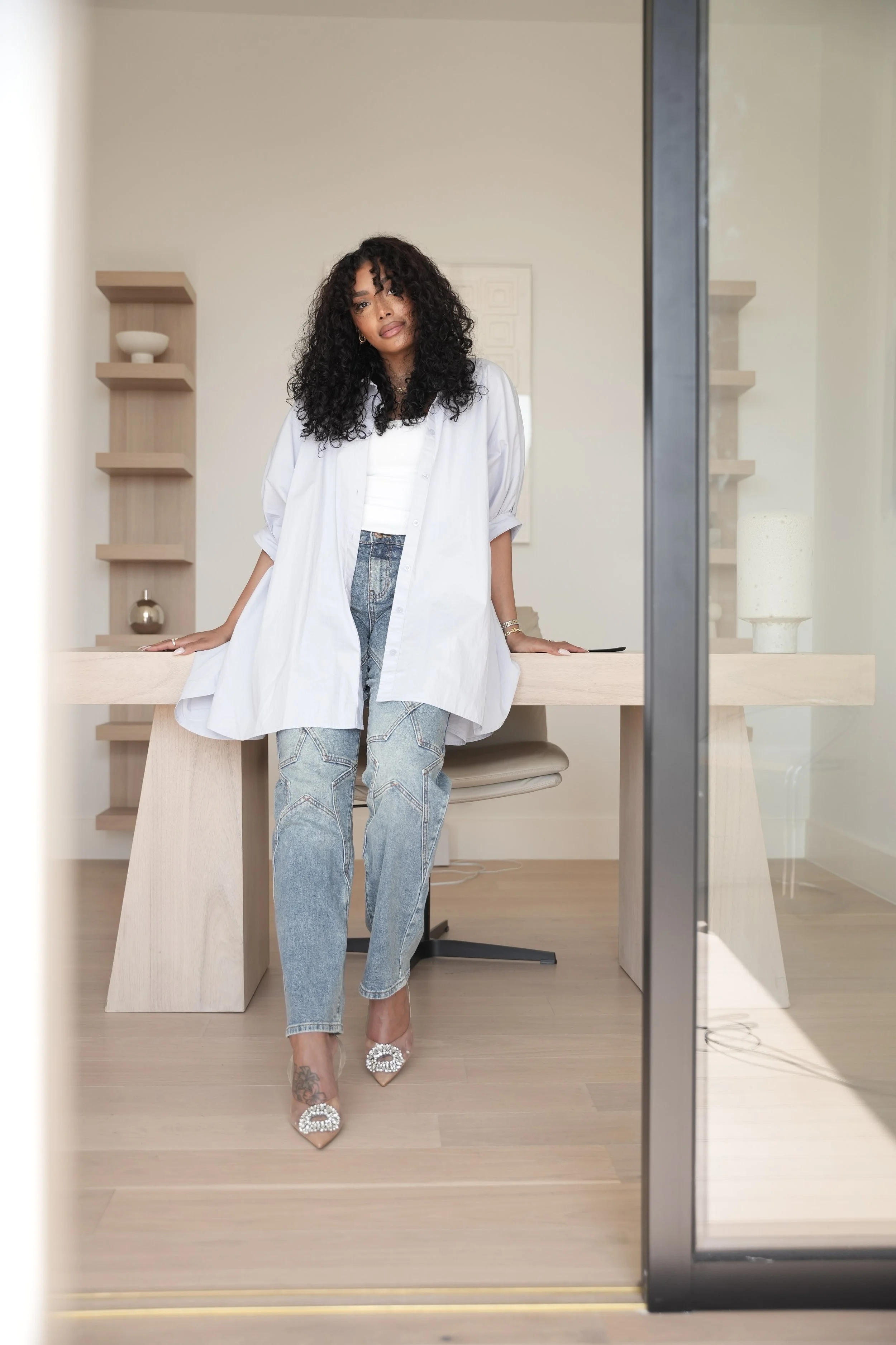 A woman with curly black hair, wearing a white shirt, light blue jeans, and embellished high heels, stands inside an office space, leaning on a wooden desk.
