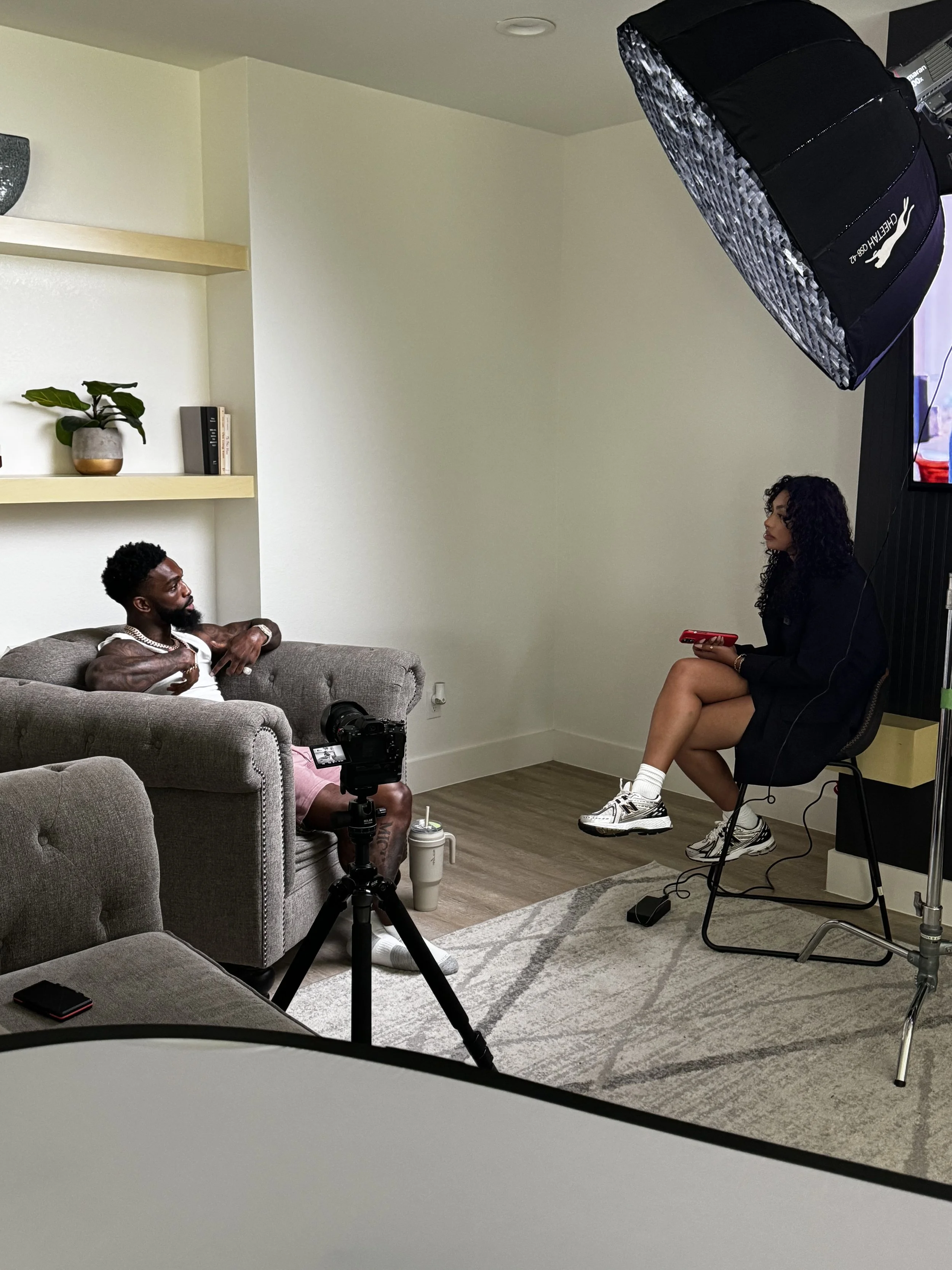 A man and a woman sitting in a room with filming equipment, including a camera on a tripod, a large lighting softbox, and a microphone, during an interview or video shoot.