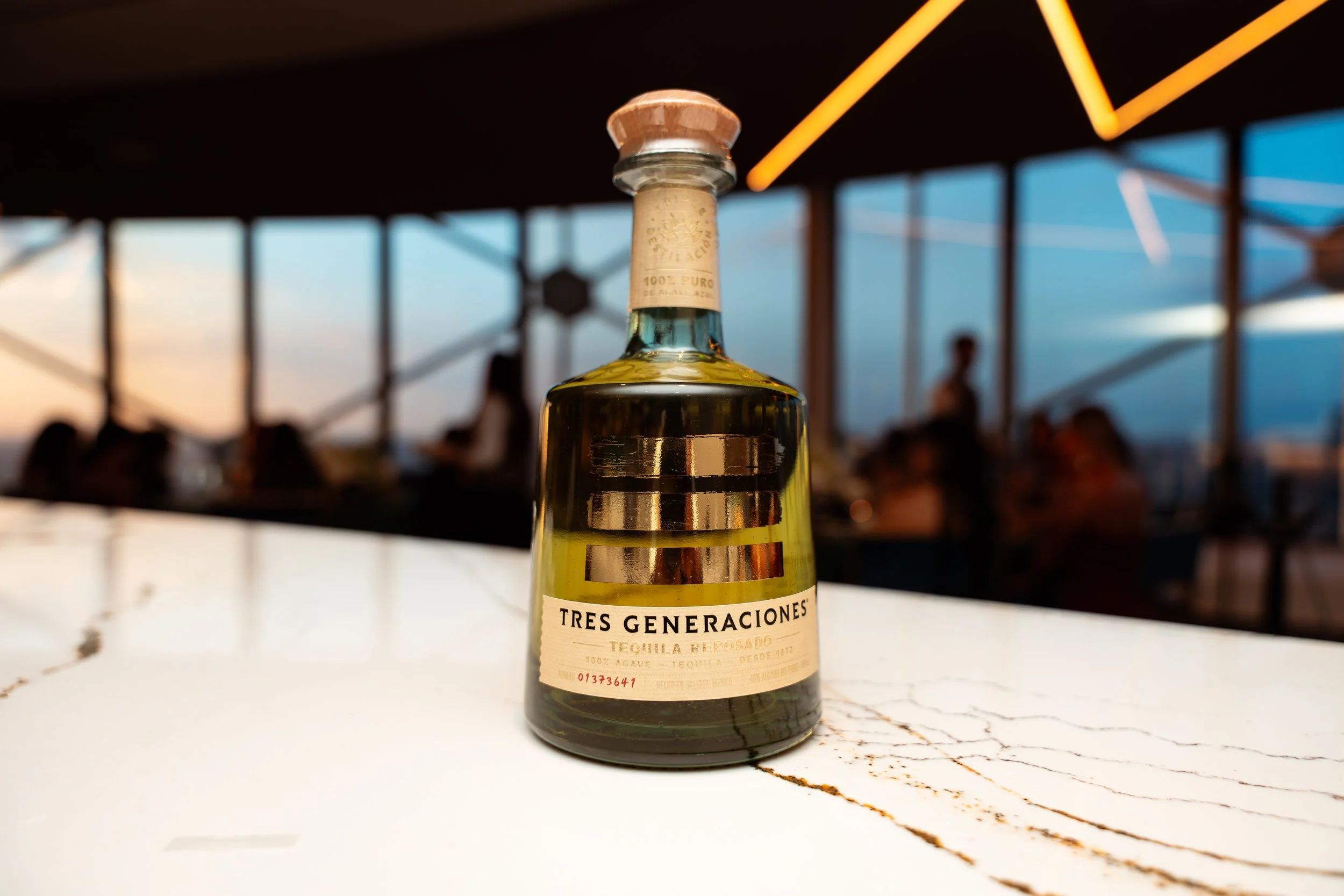 A bottle of Tres Generaciones tequila with a white label, sitting on a white surface with a cracked texture, during a sunset at an outdoor venue with people in the background and a view of the sky.