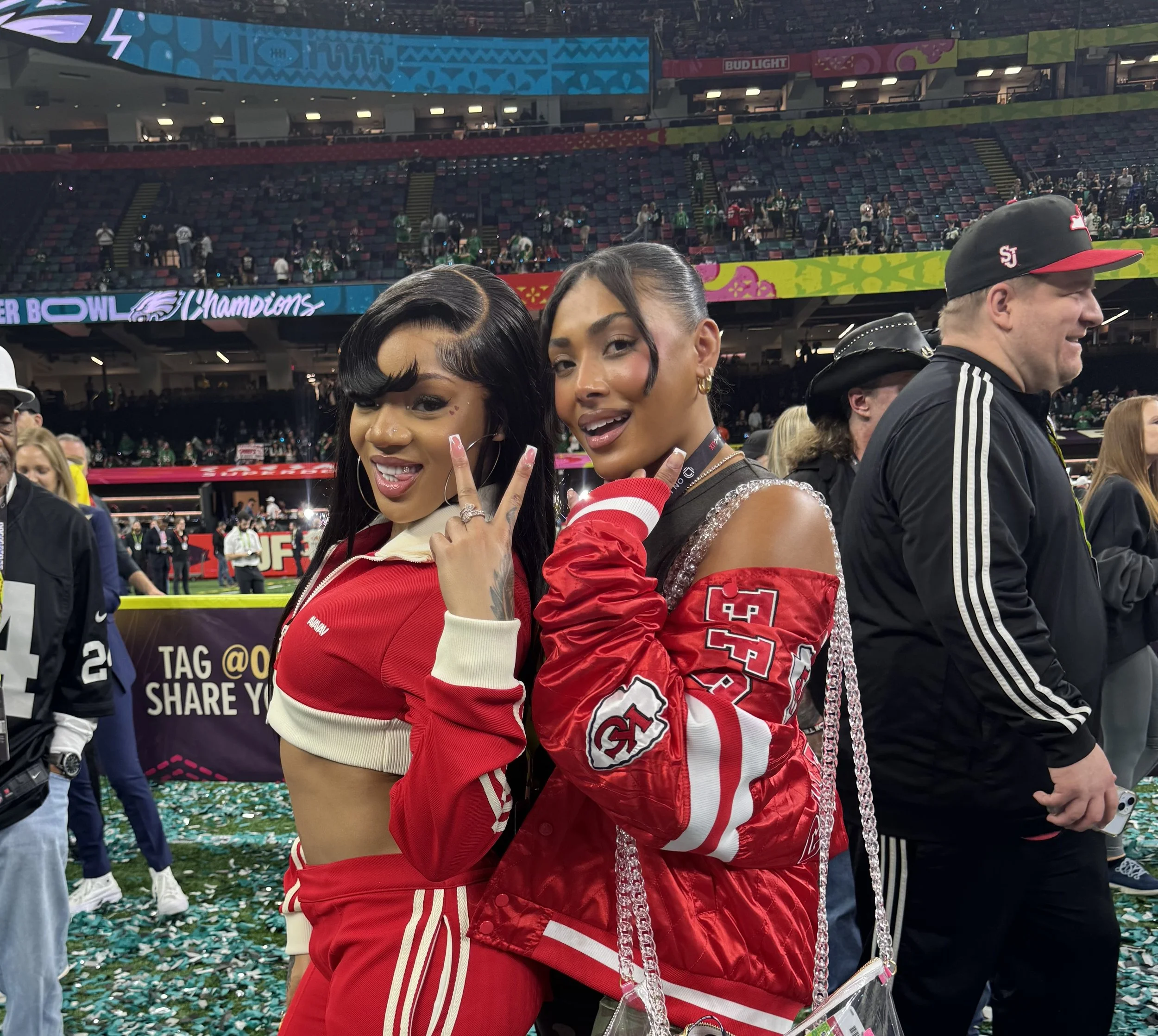 Two women celebrating at a sports event in a stadium, wearing red sports jerseys, with confetti on the ground and people in the background.