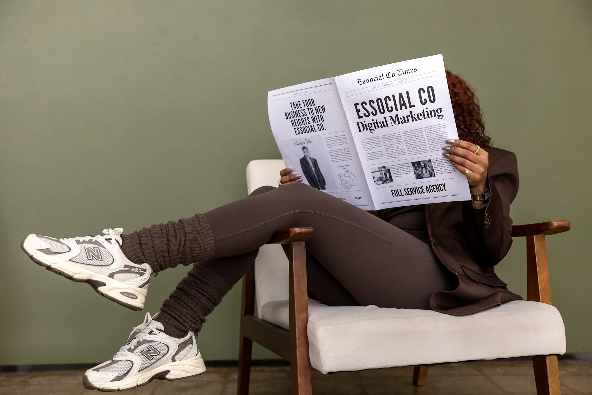 Person sitting on a cream-colored sofa, reading a newspaper, wearing brown lounge clothes and white New Balance sneakers with brown socks, against a plain green wall.