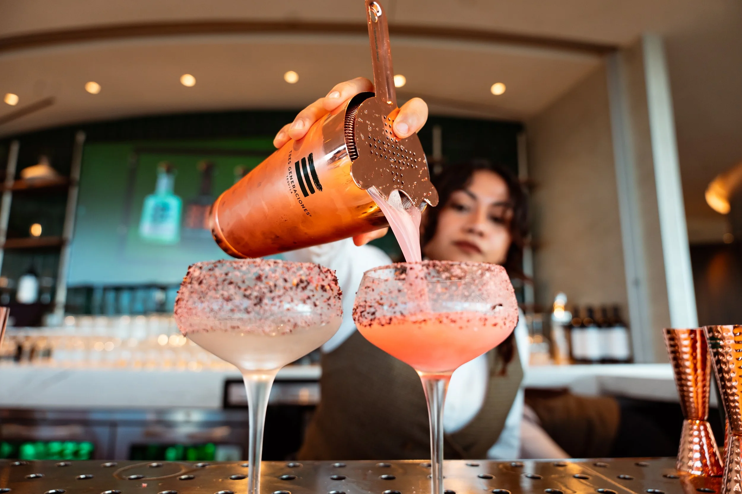 Bartender pouring a pink cocktail through a shaker into two margarita glasses with salted rims at a bar.