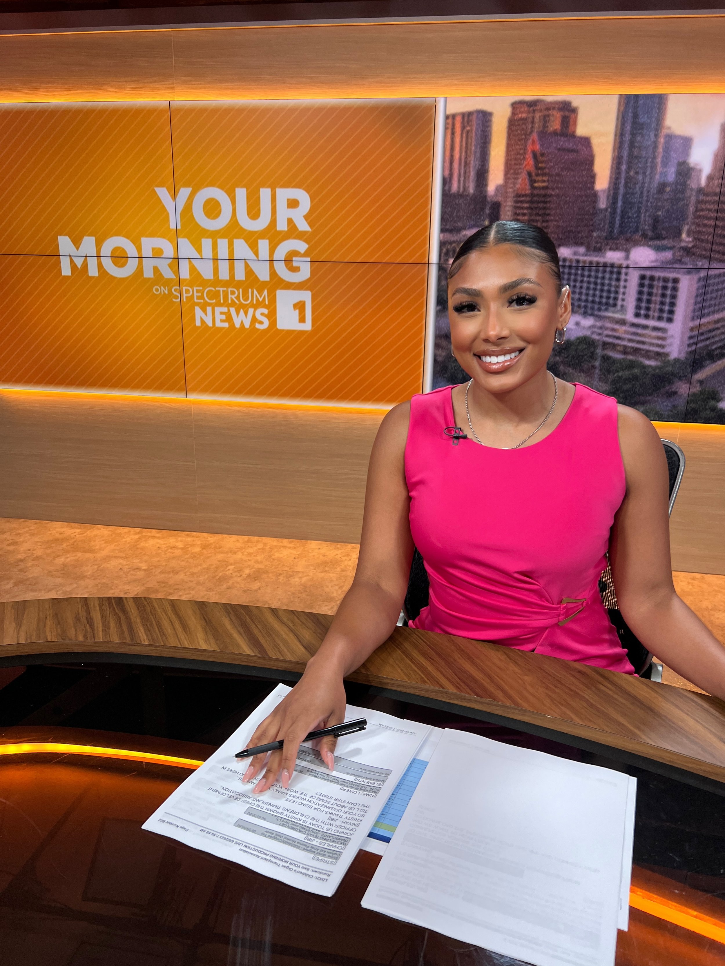 A female news anchor smiling at the camera in a television studio. She is sitting at a wooden desk with papers and a pen in front of her. Behind her is a large screen displaying the text 'Your Morning on Spectrum News 1' with a cityscape background.