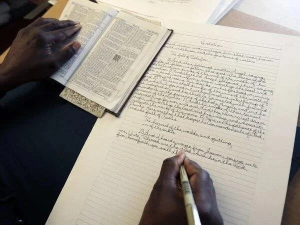 A person writing in a notebook with an open dictionary nearby, on a desk.