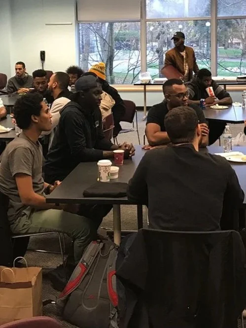 A diverse group of people sitting around tables in a room with large windows, participating in a meeting or discussion.