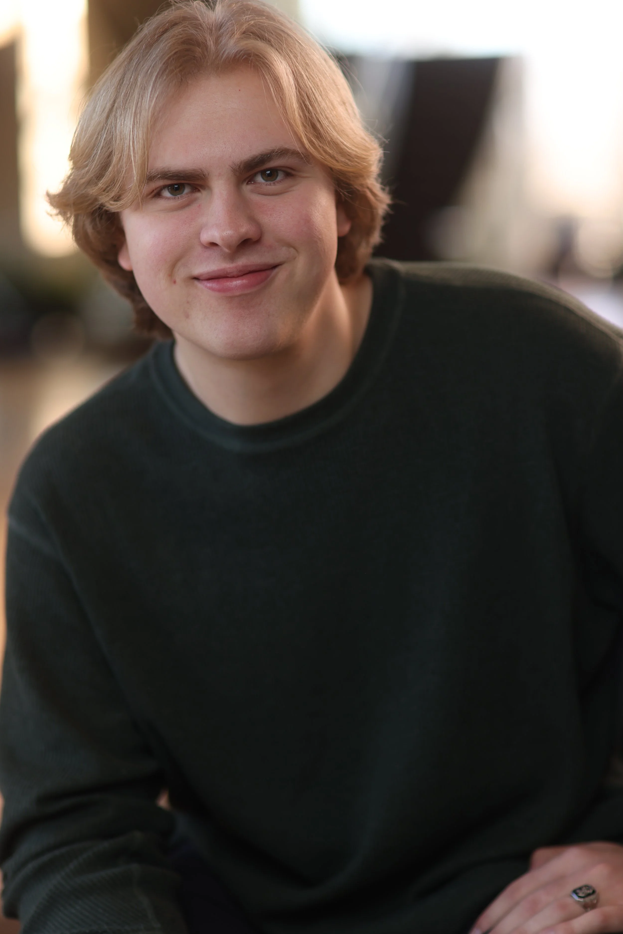 A young man with shoulder-length blond hair, wearing a dark sweater, smiling at the camera, in an indoor setting with natural light.
