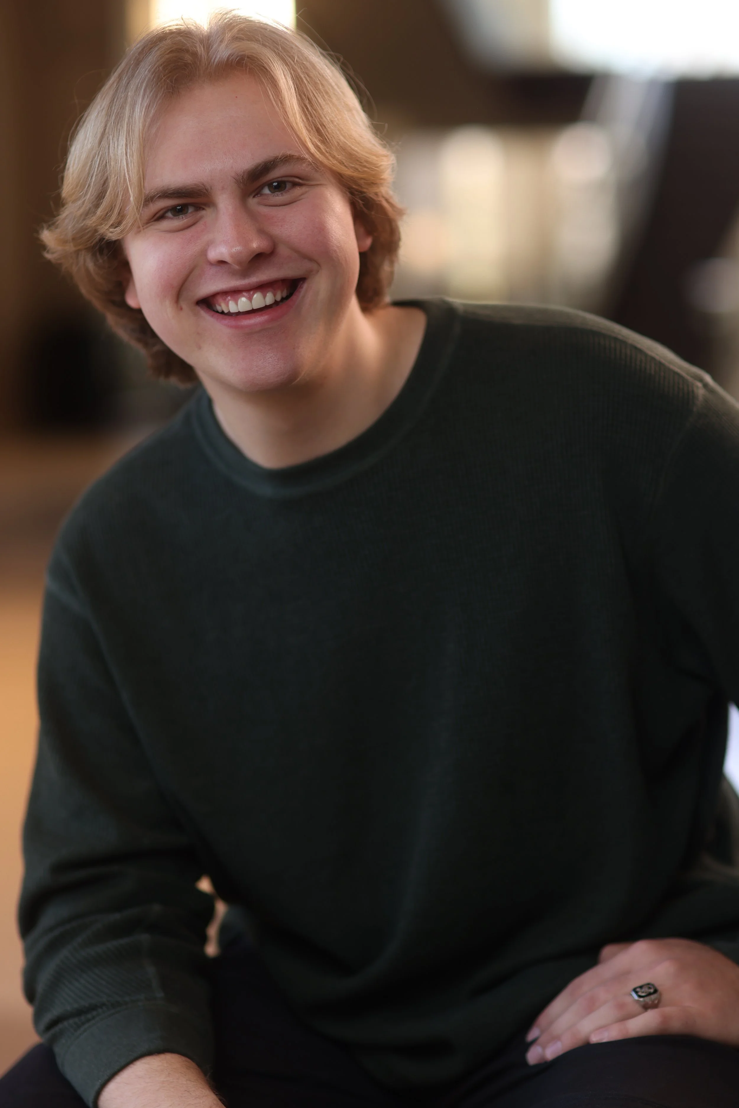 Young man with blond hair smiling, wearing a black sweatshirt, sitting indoors with a blurred background.