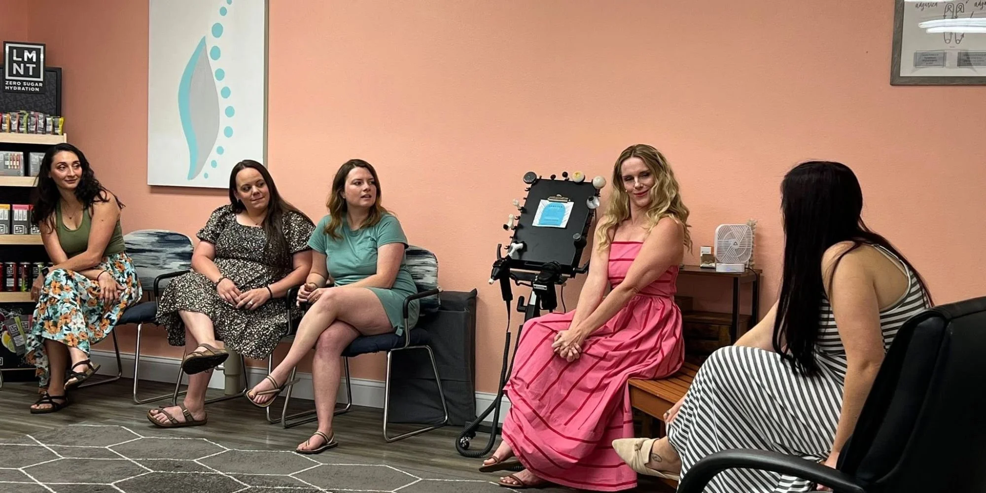 Five women sitting and engaging in a conversation in a room with pink walls. One woman in a pink dress is sitting on a bench, smiling, and next to a camera on a tripod. The others are seated on chairs, wearing casual dresses, with bookshelves in the 