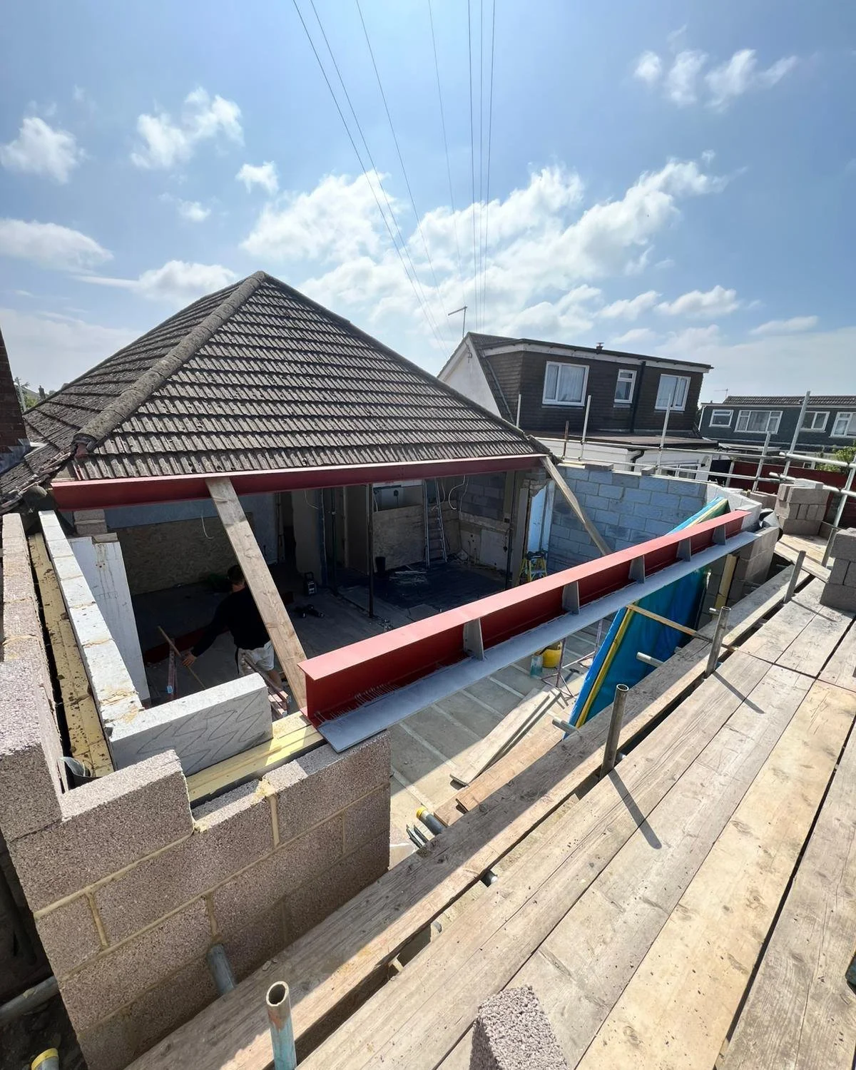 Construction site showing a partially built house with workers installing a roof, surrounded by scaffolding and building materials under a partly cloudy sky.