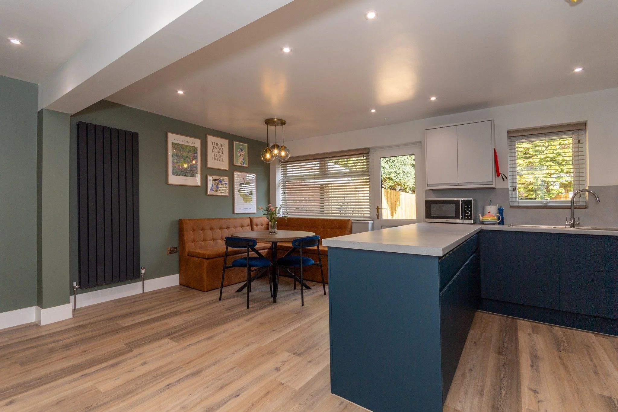 Open-concept kitchen and dining area with wood flooring, green accent wall, and a corner built-in dining bench with a round table and seating. Modern decor includes a gallery wall, pendant lights, and a window with blinds.