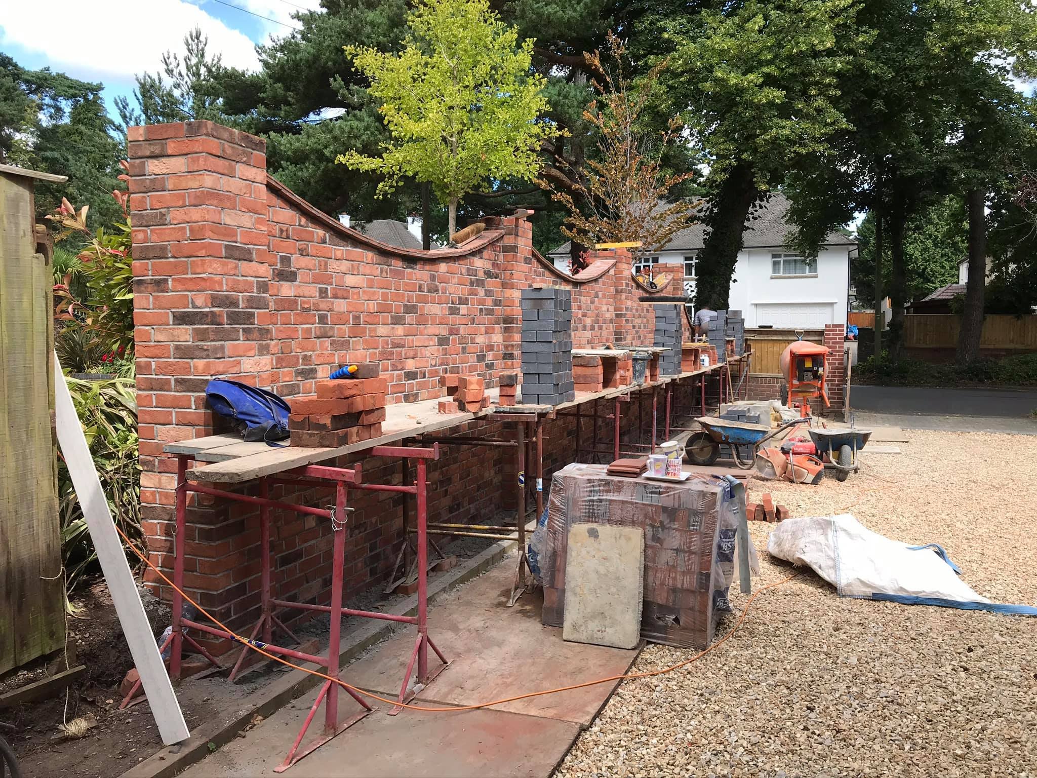 Construction site with partially built brick wall, scaffolding, construction tools, and materials in a residential yard.