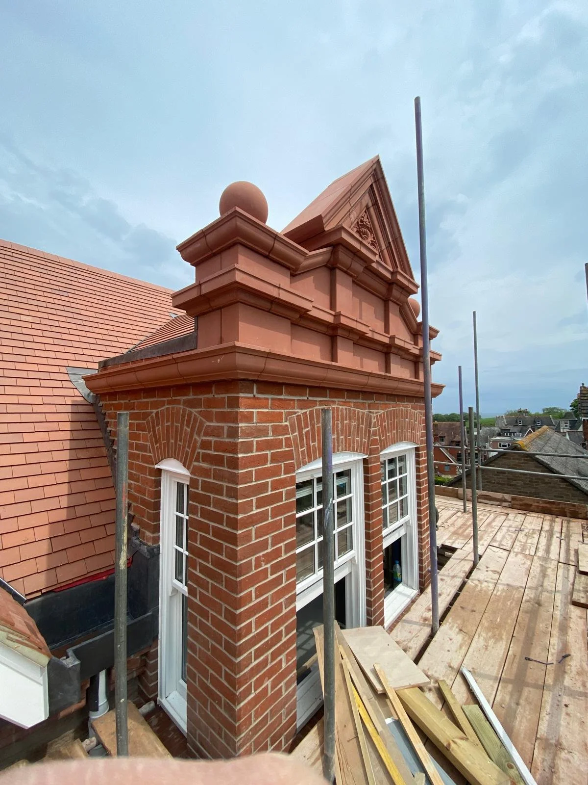 View of a rooftop under construction showing a brick chimney with decorative top and scaffolding, with neighboring rooftops and a cloudy sky in the background.