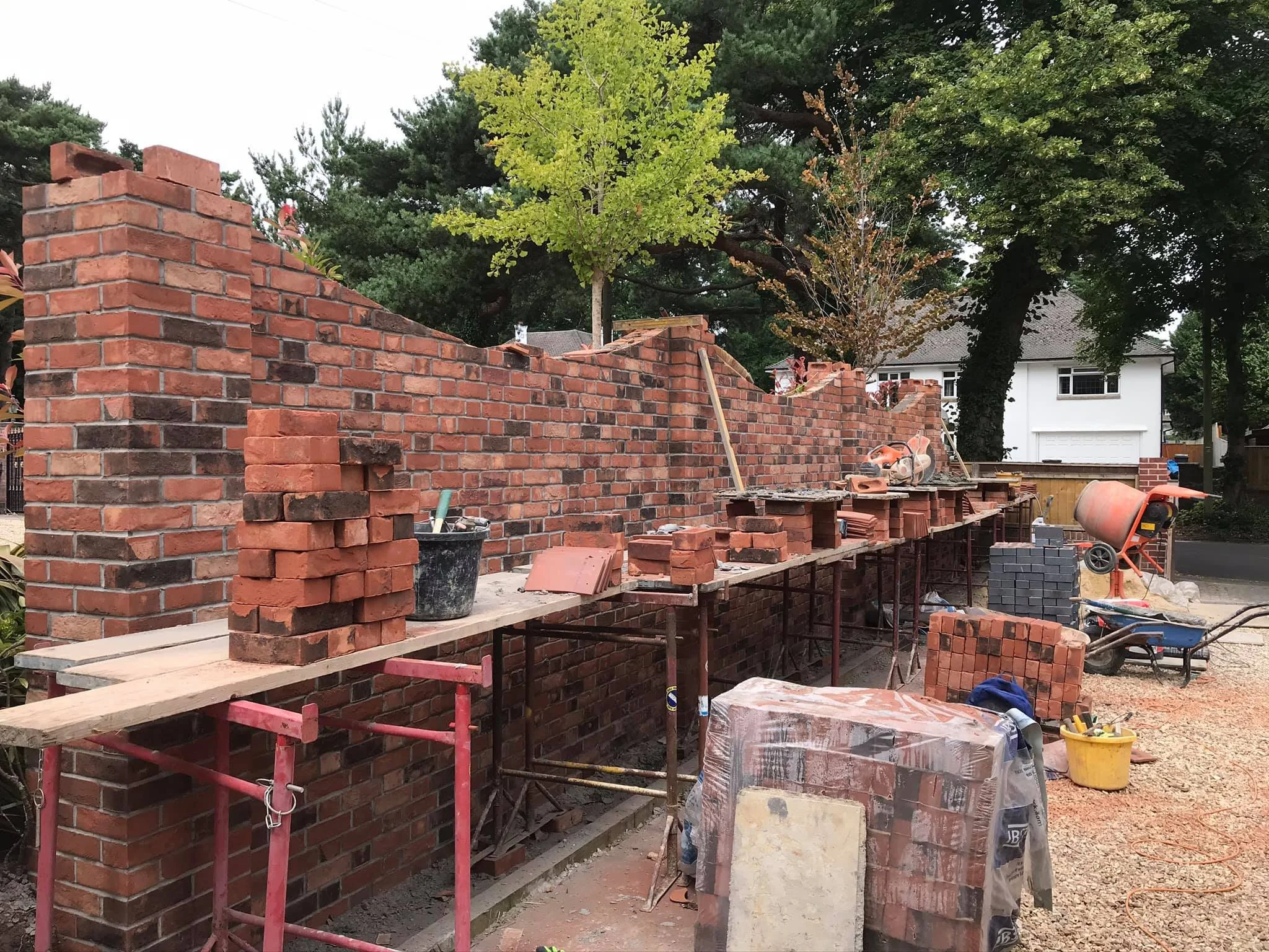 Construction site with a brick wall being built, construction tools, bricks, and a cement mixer, with trees and houses in the background.
