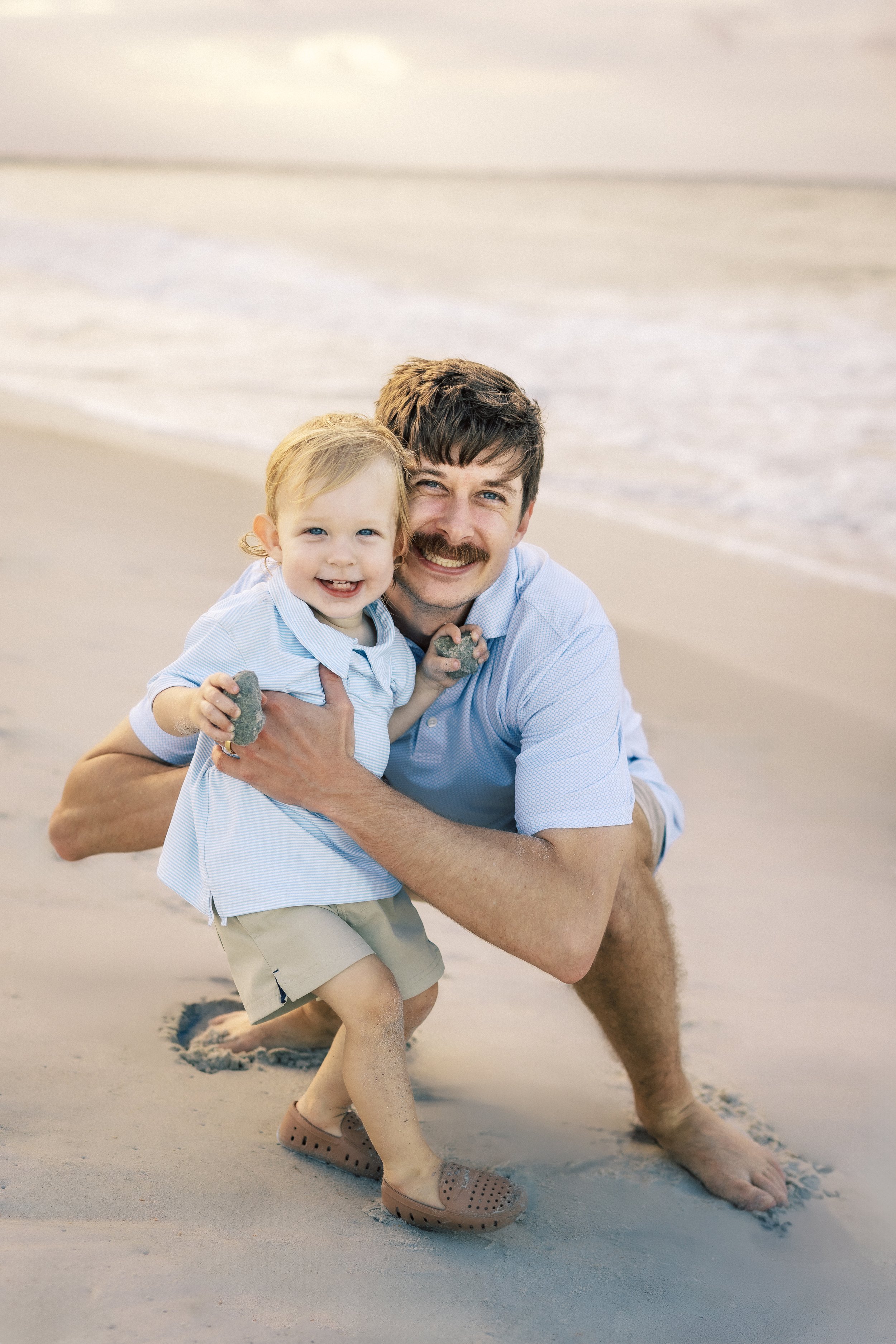 A man with a mustache and a young boy with blonde hair smiling at the camera on the beach, with waves in the background. They are squatting in the sand, holding small rocks, and smiling. Photographer Amelia Island, Jacksonville, St Augustine, St Simo