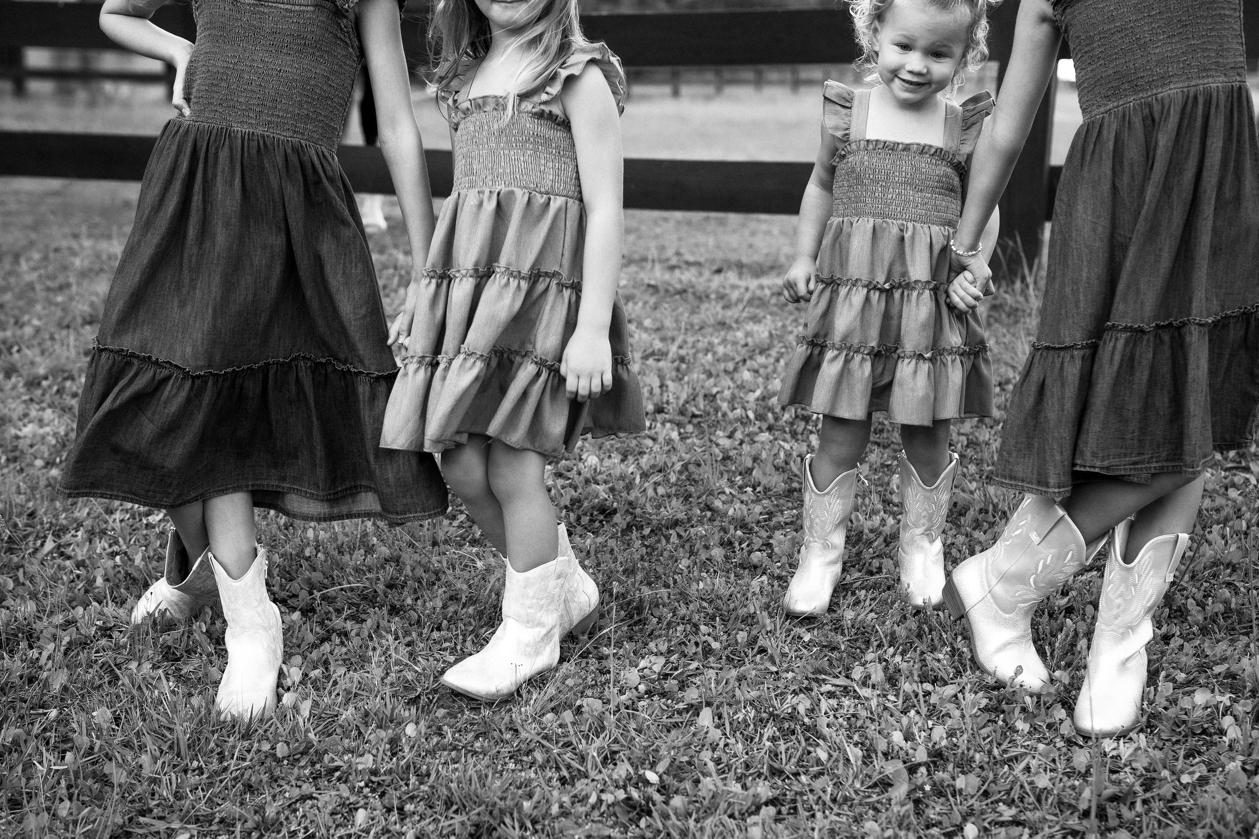 Four children standing outdoors in a grassy area, dressed in similar flowy dresses and cowboy boots, with their legs close together, facing the camera.